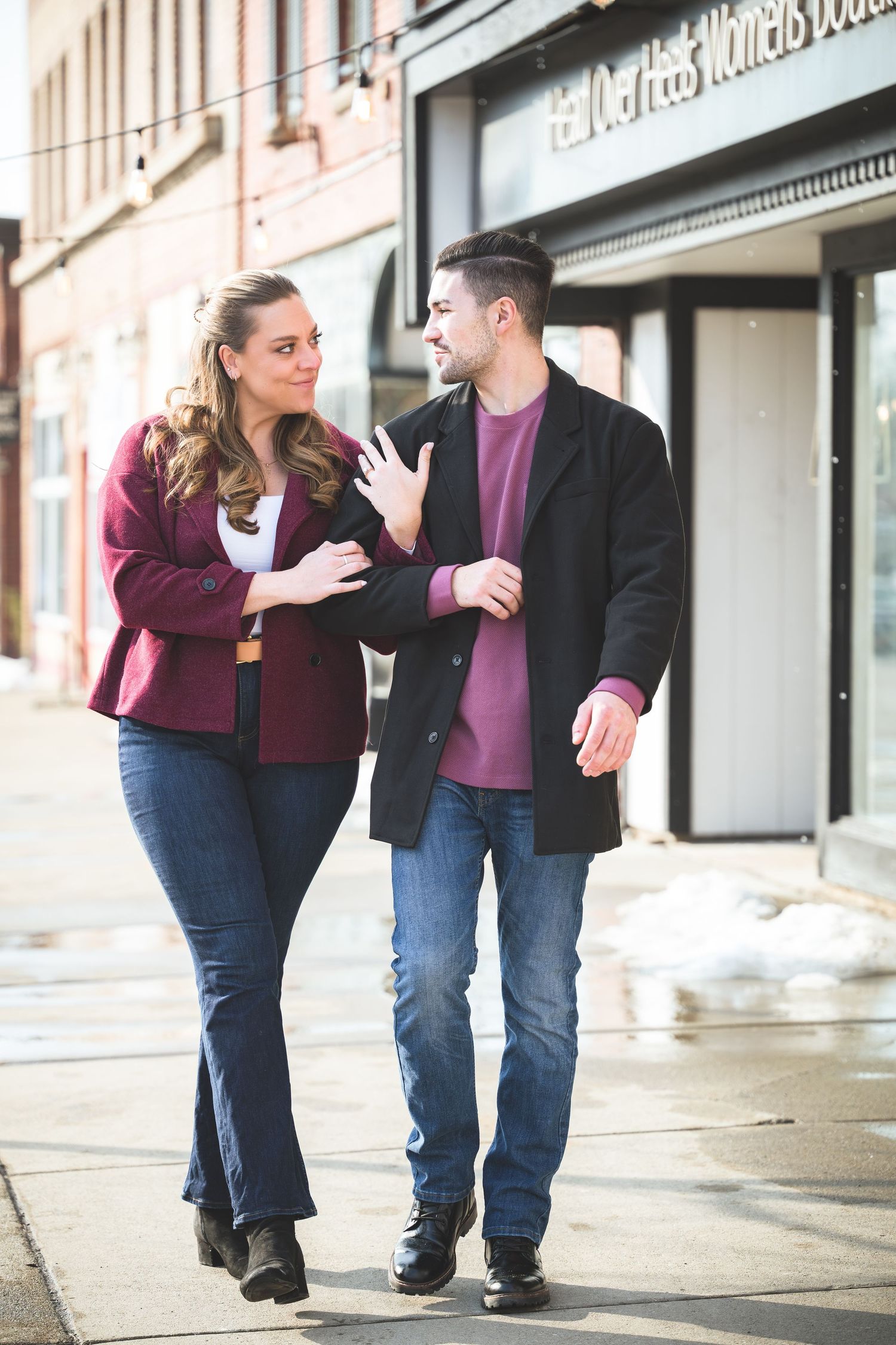 Happy couple walking together on a sunny winter sidewalk, both wearing burgundy and black outfits.