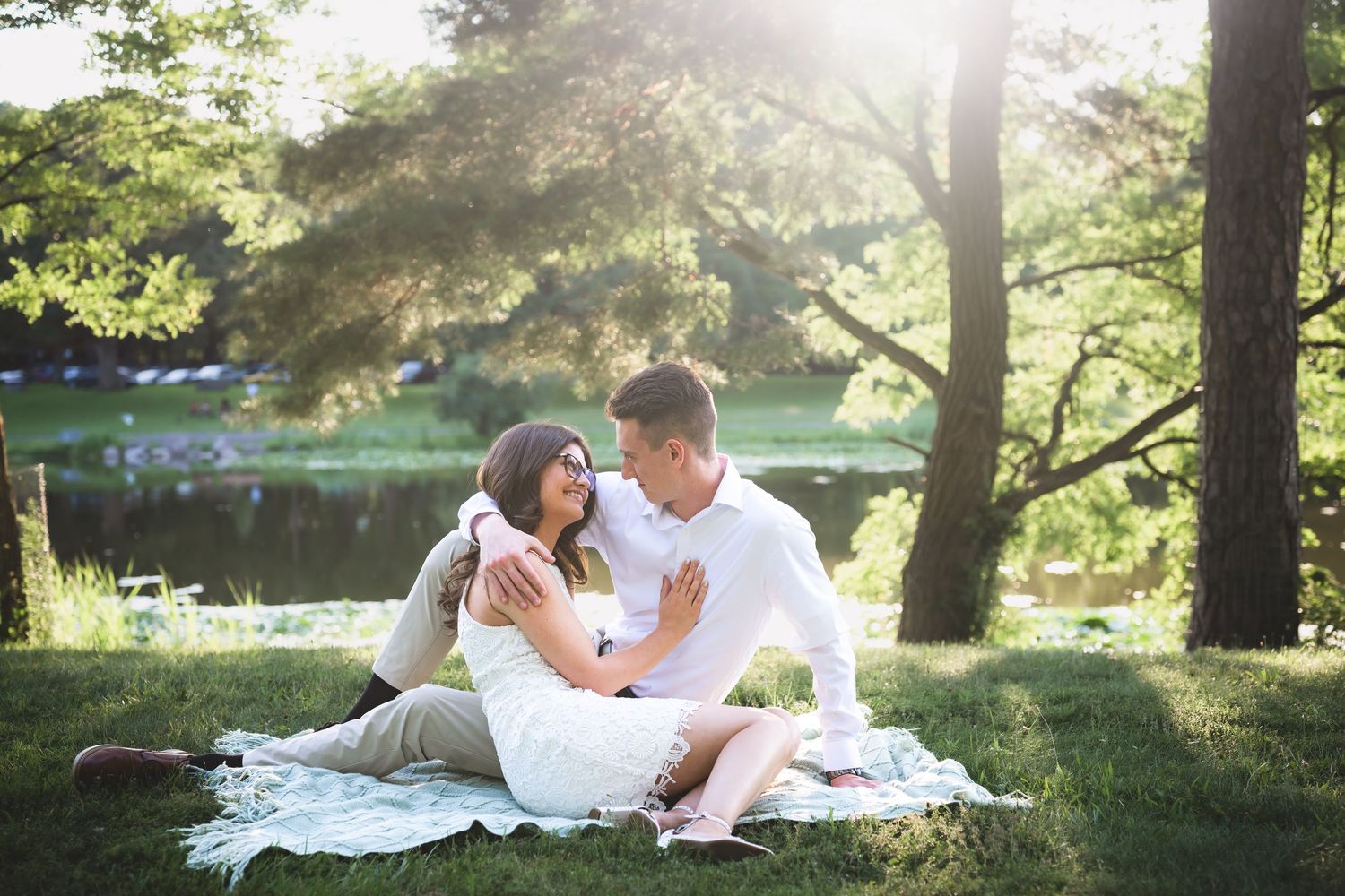 Couple sitting on a blanket in a sunny park, sharing a romantic moment near a serene lake surrounded by lush green trees.