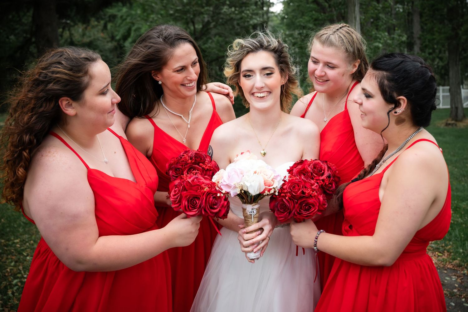 Bride in white dress with bridesmaids in red dresses holding vibrant red bouquets in outdoor garden setting.