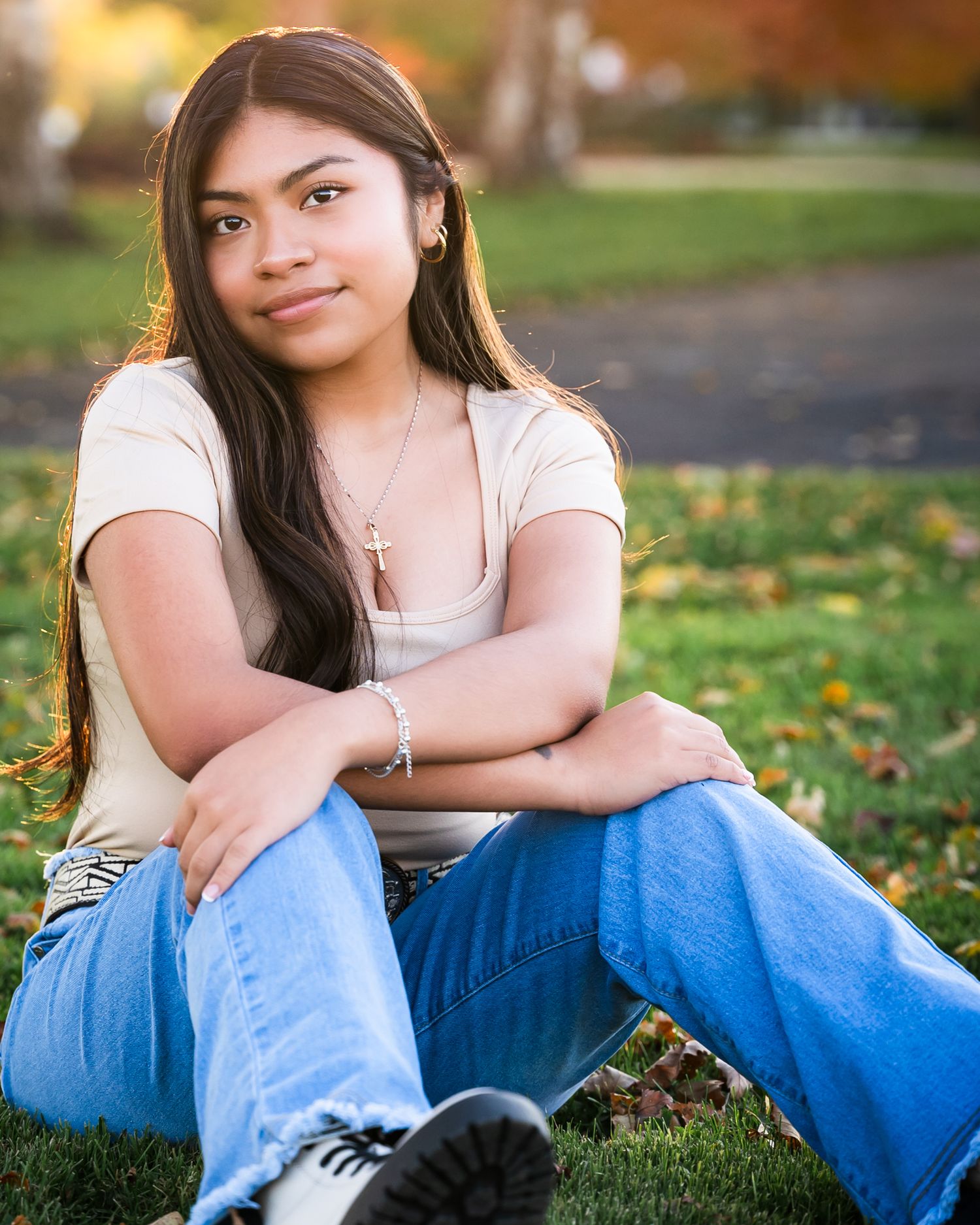 A person in blue jeans sits on grass with autumn leaves during a casual outdoor photo session.
