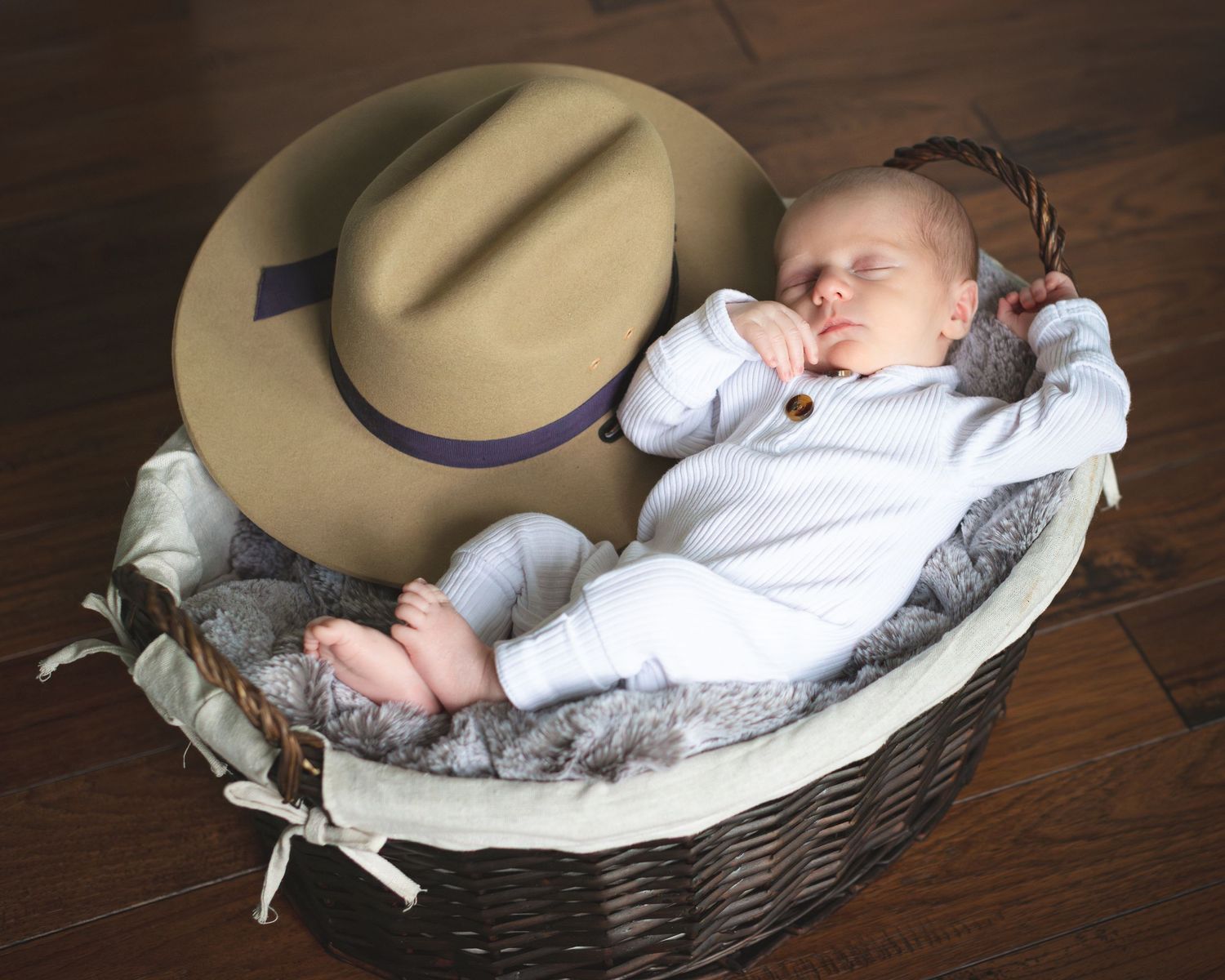 A sleeping baby lies in a woven basket with a beige hat and white blanket against a dark wooden background.