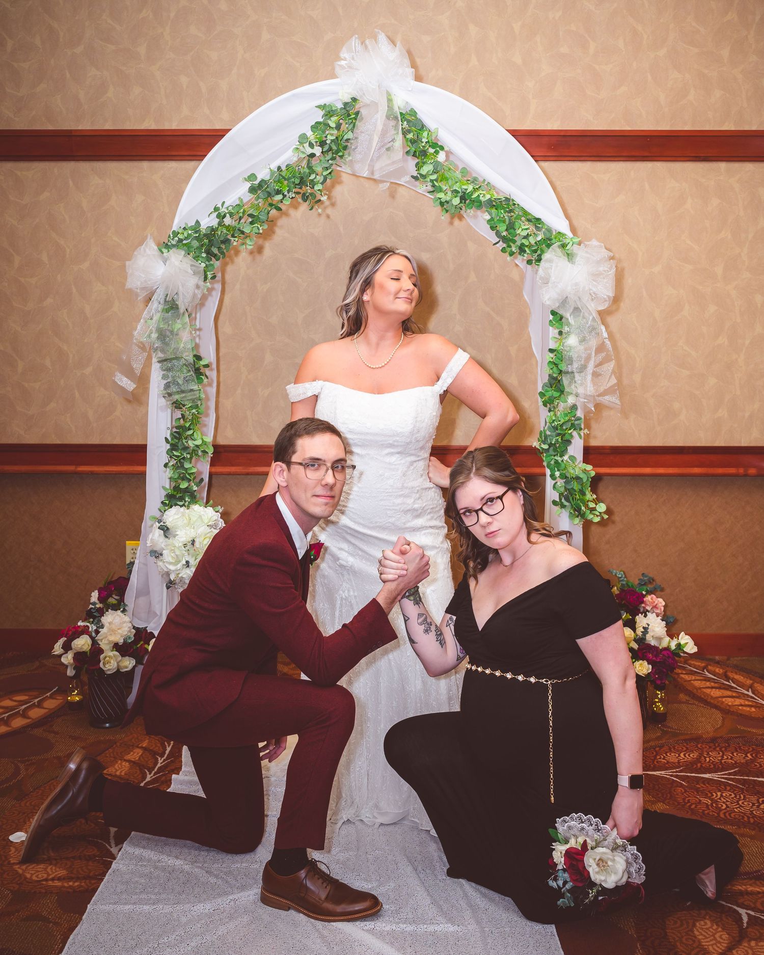 Playful wedding photo with three people posing under a decorated white arch with greenery.