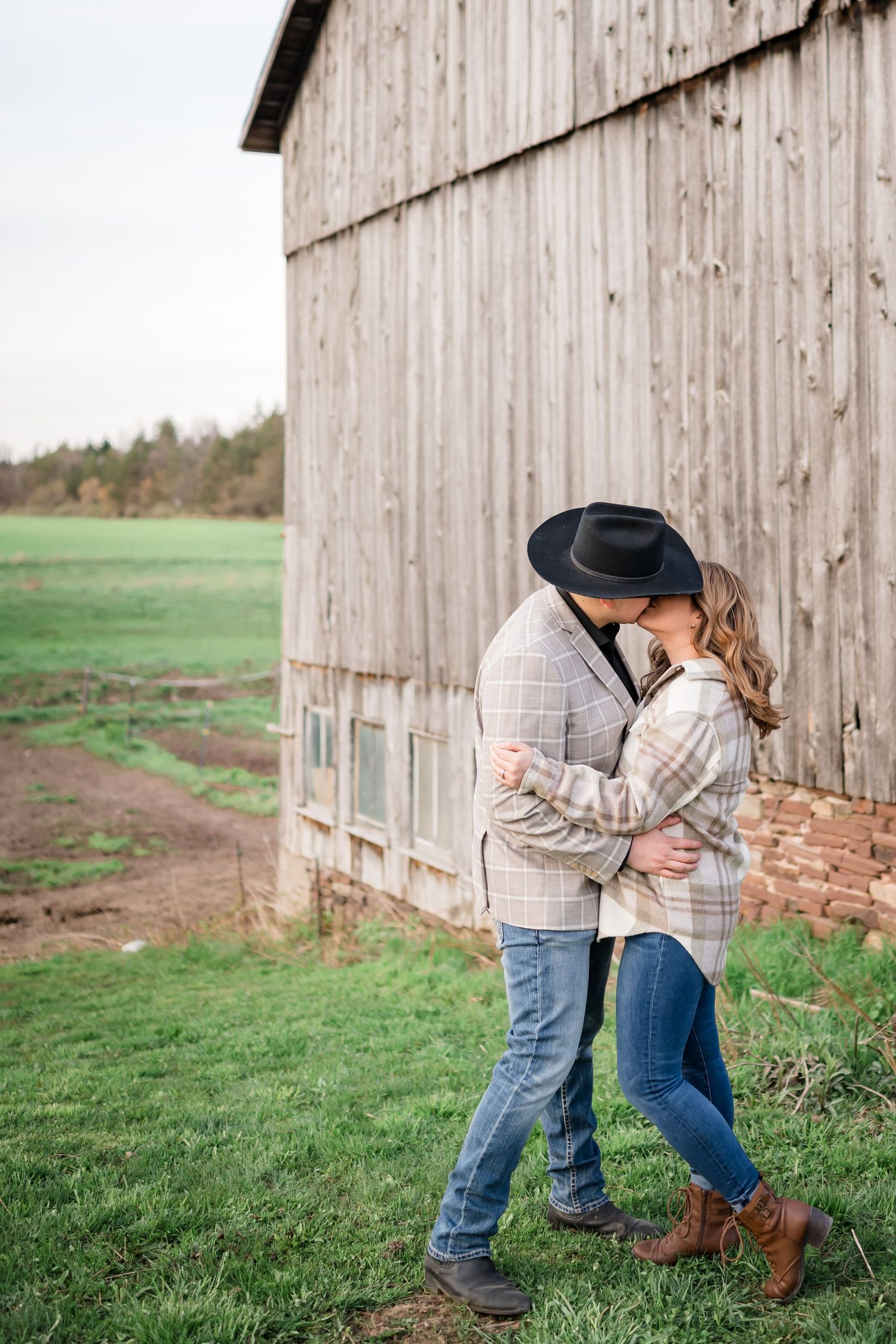Romantic couple embracing beside rustic barn wall in countryside setting wearing matching plaid jackets.