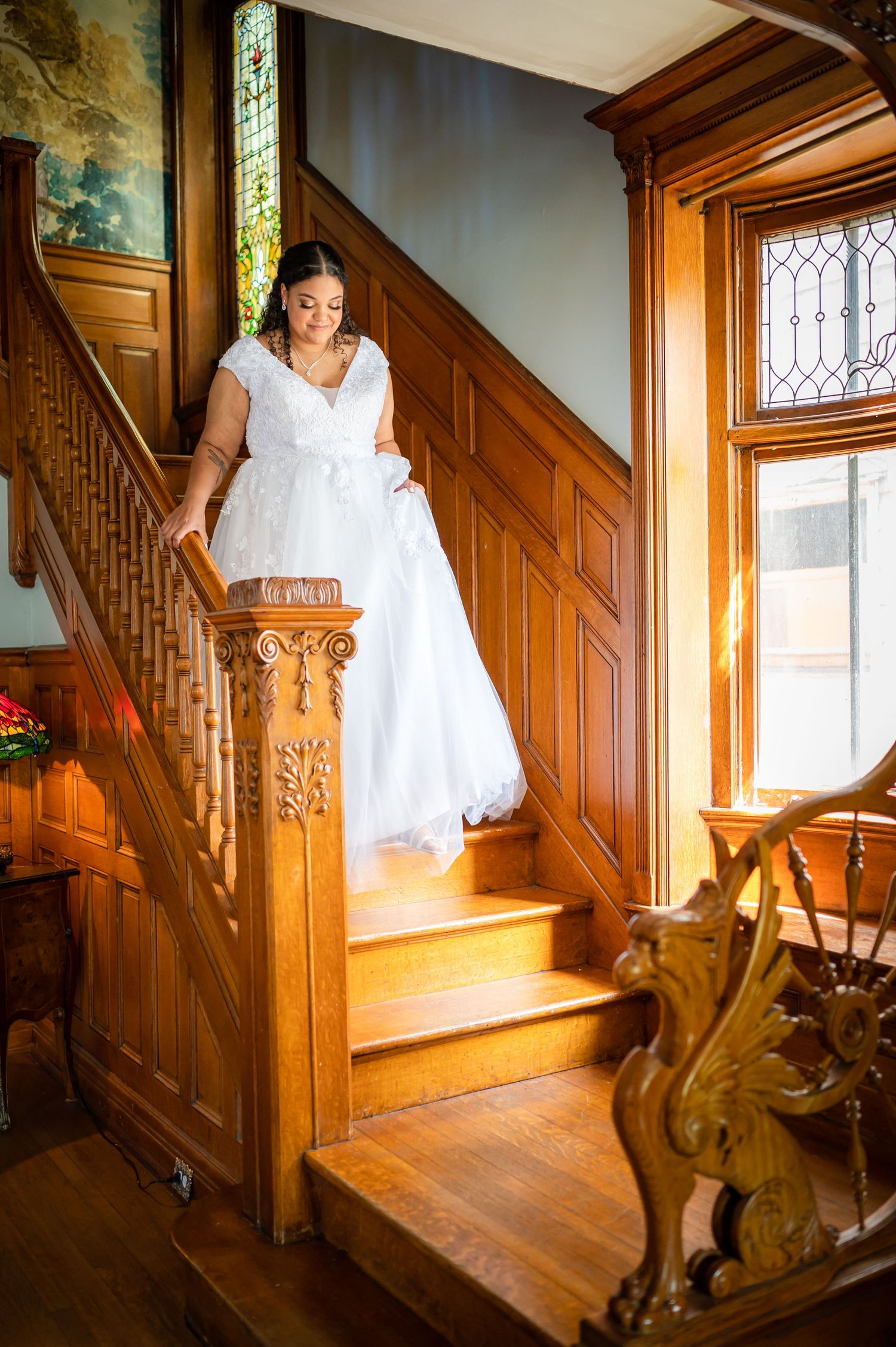 A person in a white wedding dress descends an ornate wooden staircase in vintage-style interior.