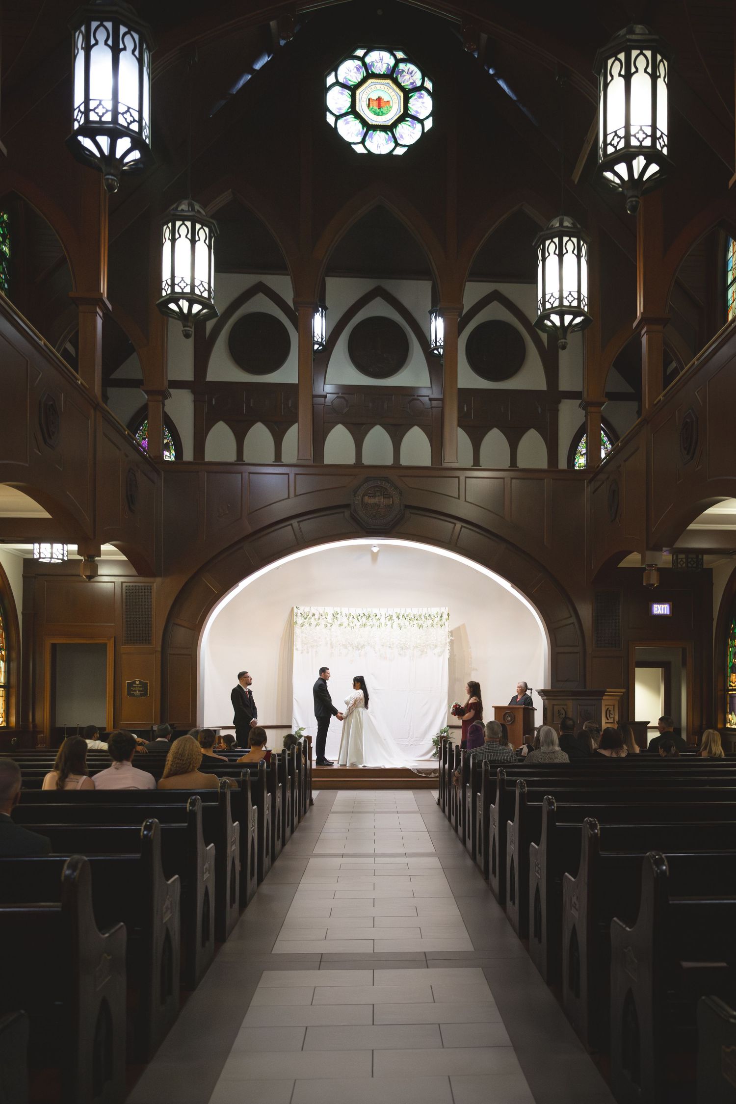 Dark wooden church interior with gothic arches and hanging pendant lights creating dramatic shadows down center aisle.