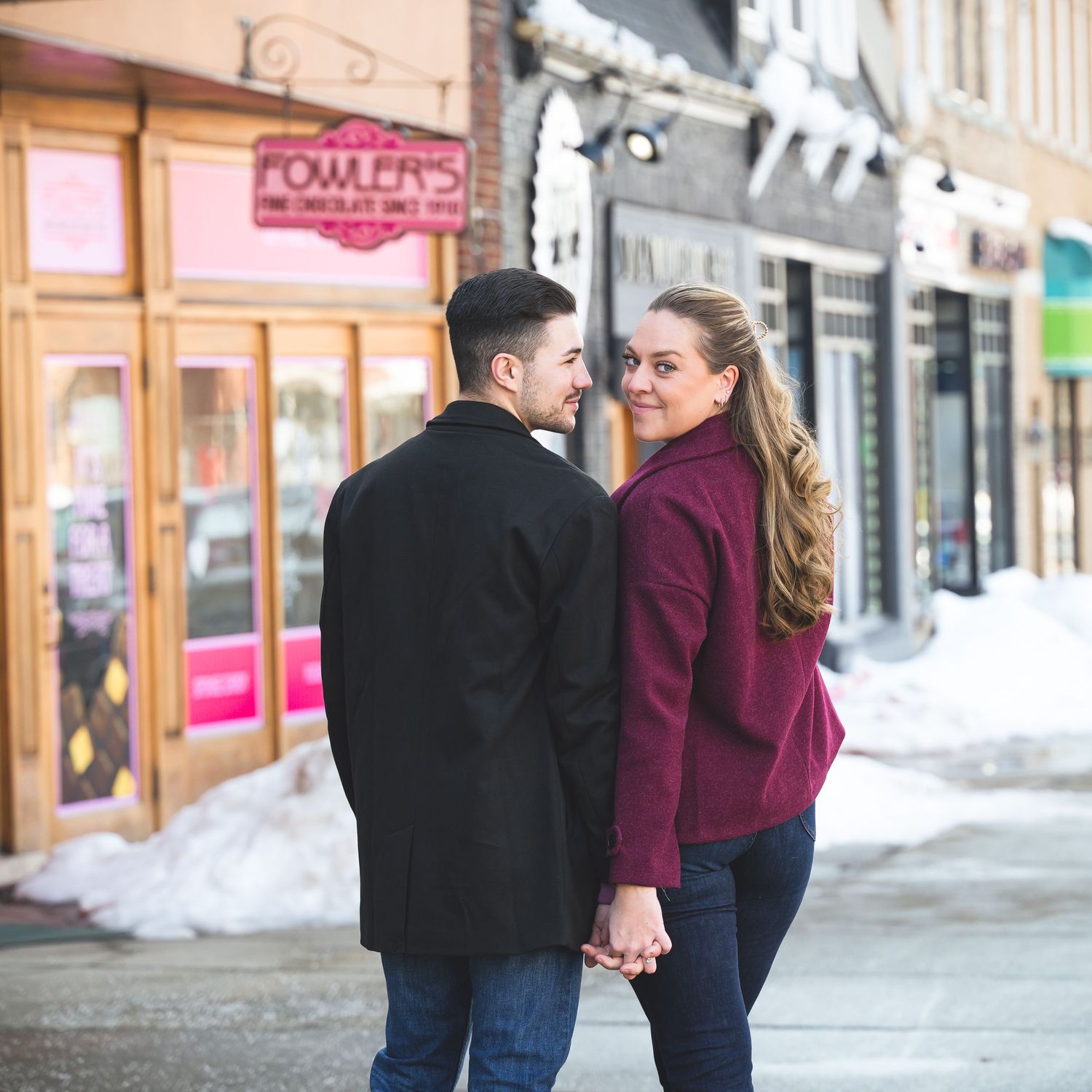 A couple holds hands on a snowy urban street, smiling at each other near a chocolate shop.