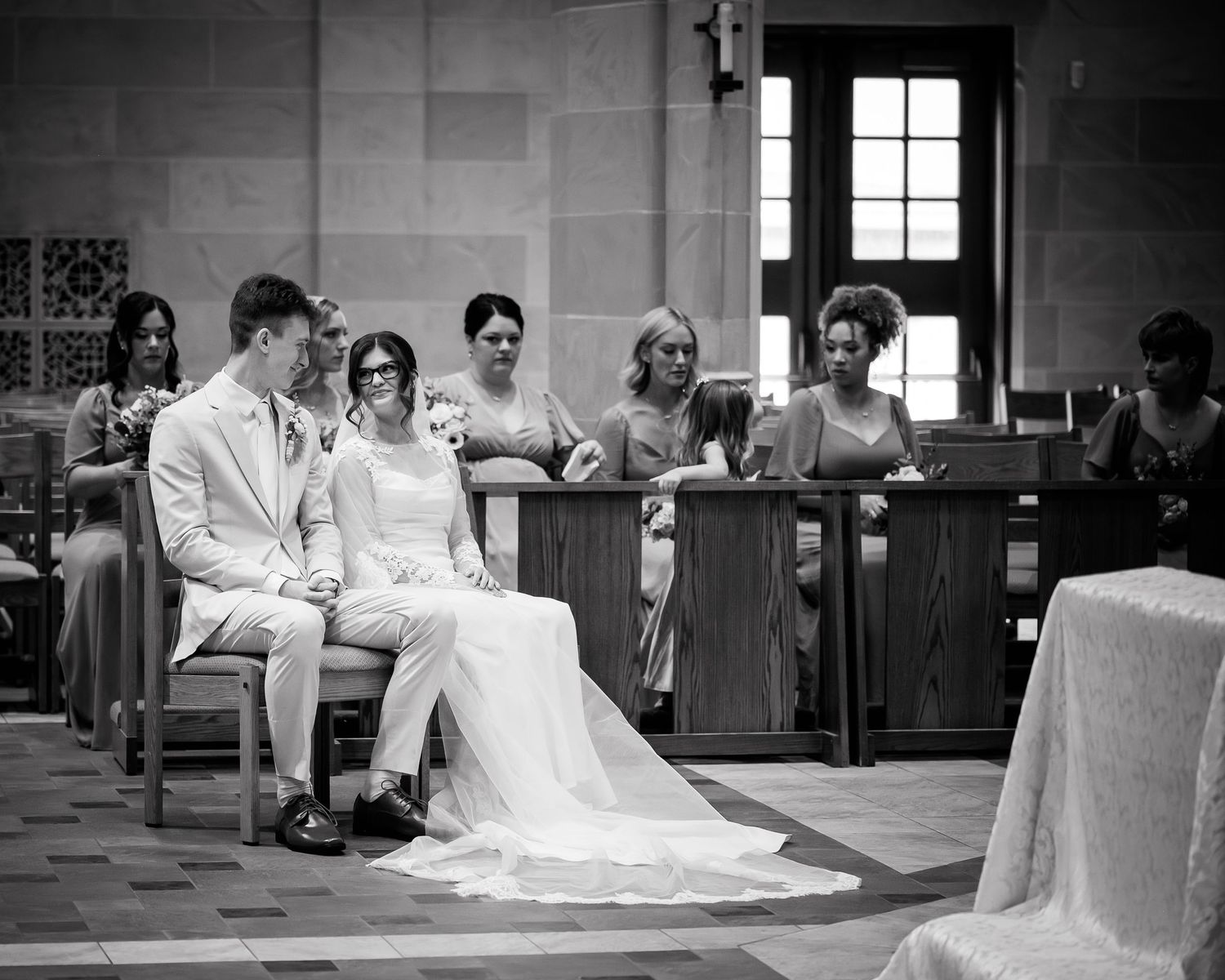 Black and white wedding photo of bride and groom seated at altar with bridesmaids in background.