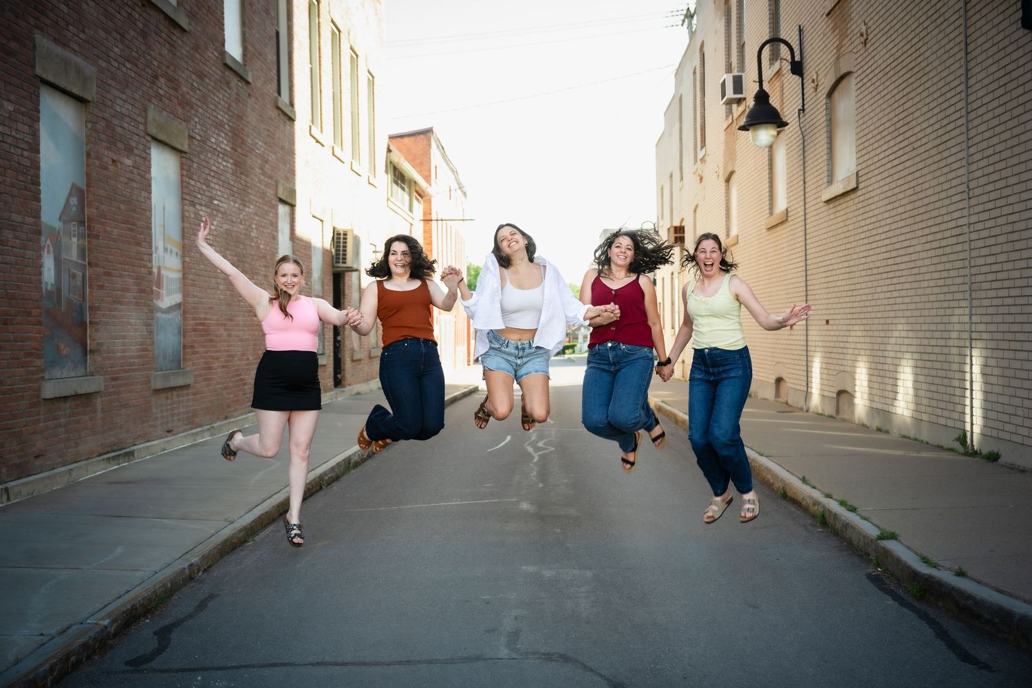 Five young women jumping joyfully in an urban alley between brick buildings.