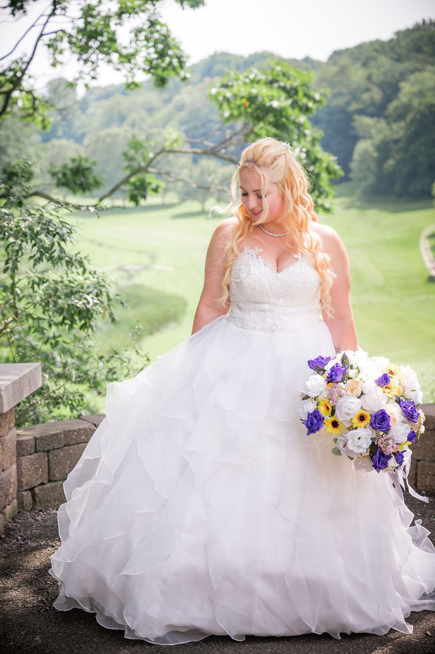 Elegant bride in layered tulle gown holding purple and yellow bouquet in outdoor garden setting.