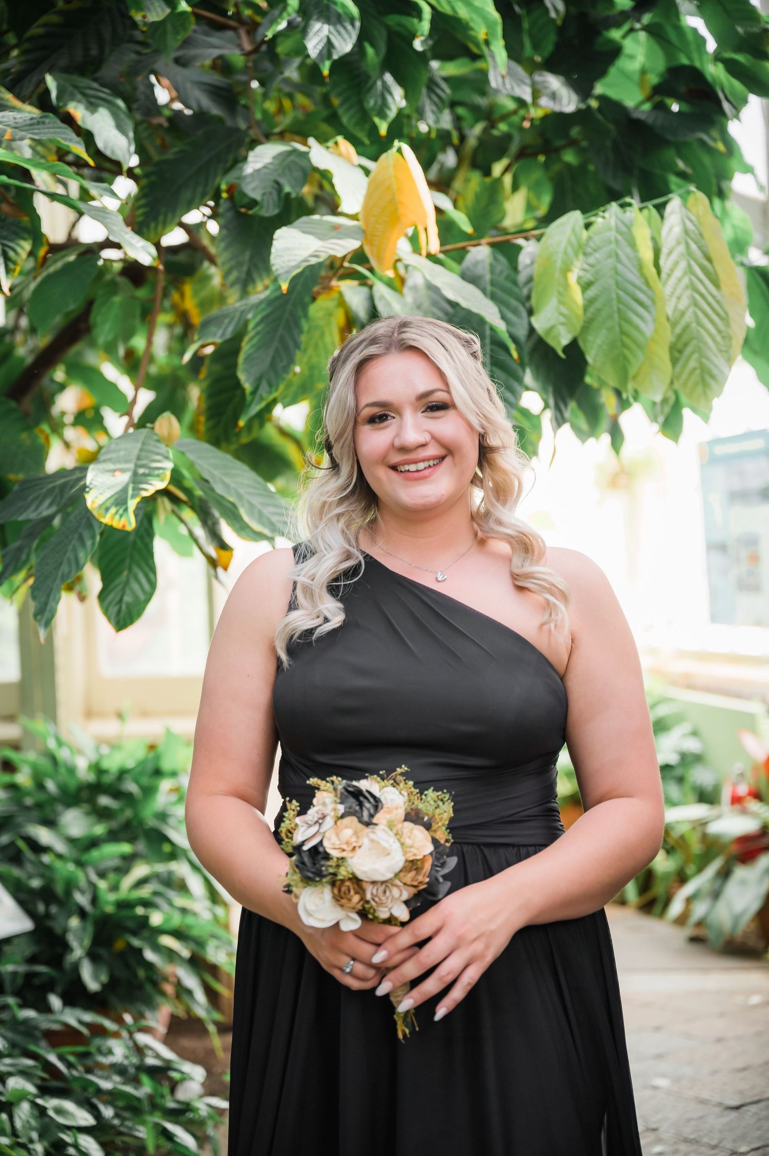 A bridesmaid in a black formal dress holds a bouquet while posing under green foliage.