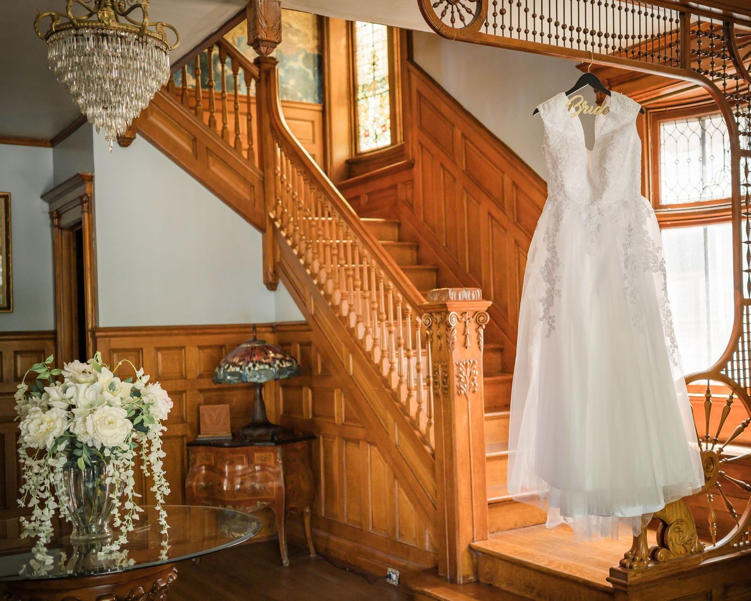 White wedding dress hanging in sunlit room with wooden staircase and crystal chandelier.