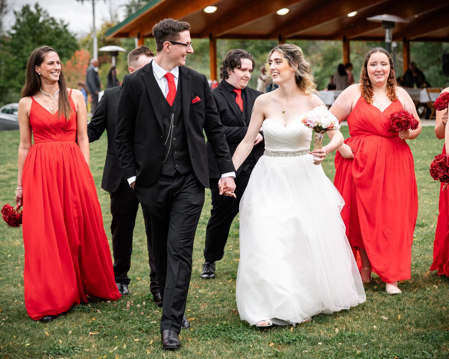 Wedding party walks on grass wearing black suits and vibrant red bridesmaid dresses.