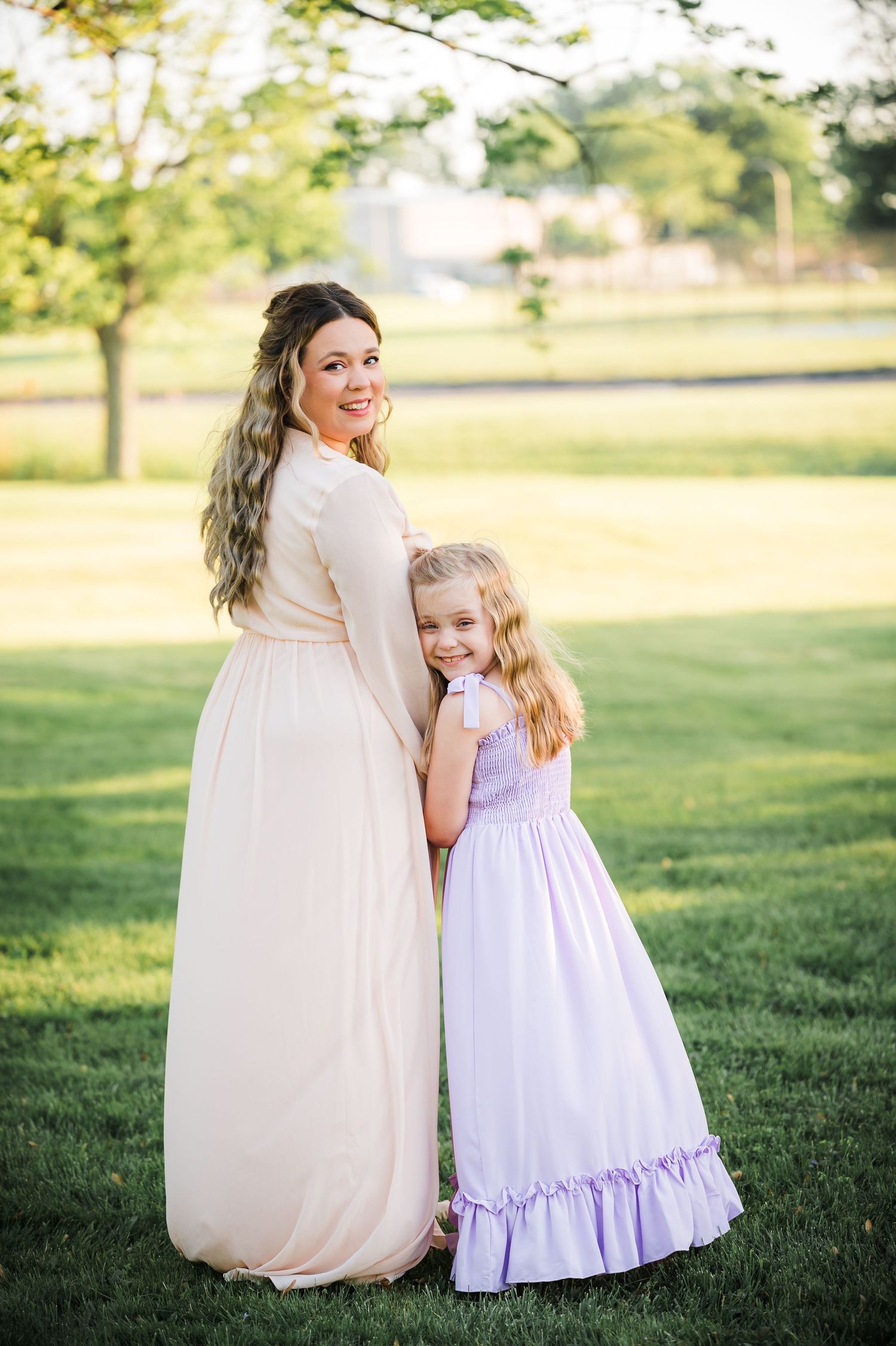 A pair in flowing dresses stand together on a sunny lawn, with the smaller one in lavender hugging close to the taller one in cream.