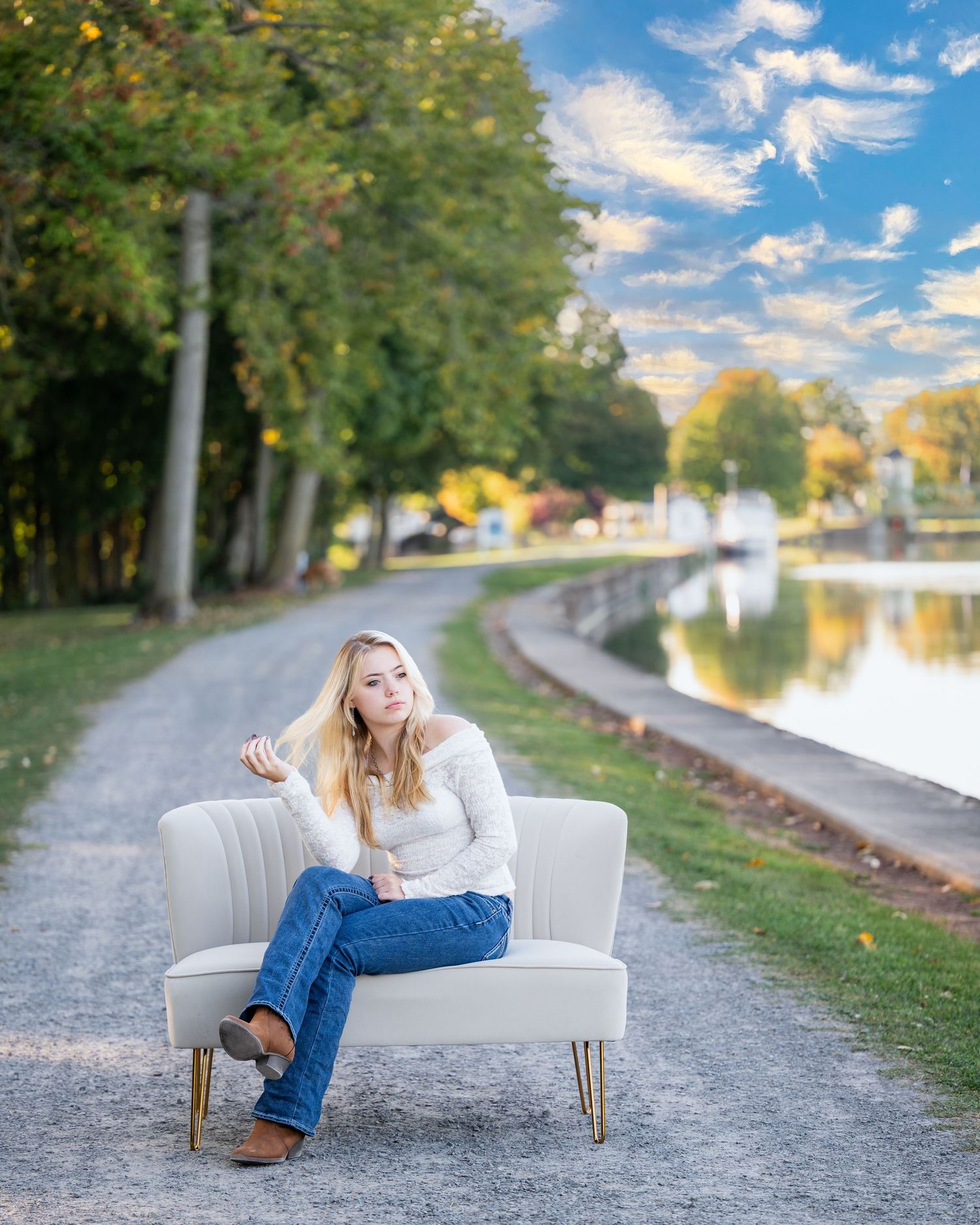 A person lounges on a white couch along a scenic canal path surrounded by autumn trees and calm water.
