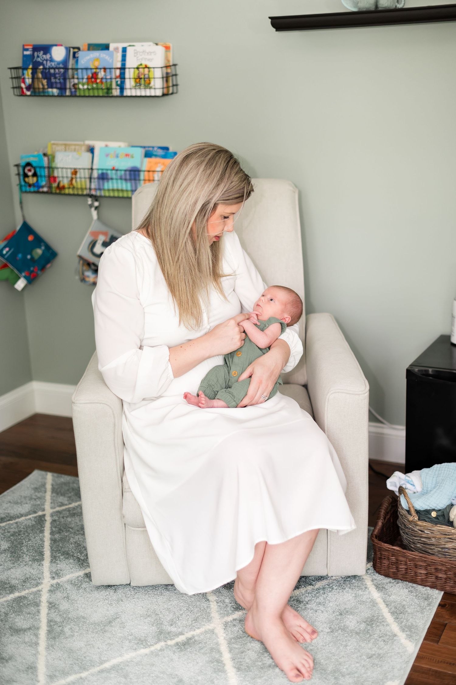 A tender moment in a nursery as a caregiver in white dress bottle feeds an infant in a comfortable armchair.
