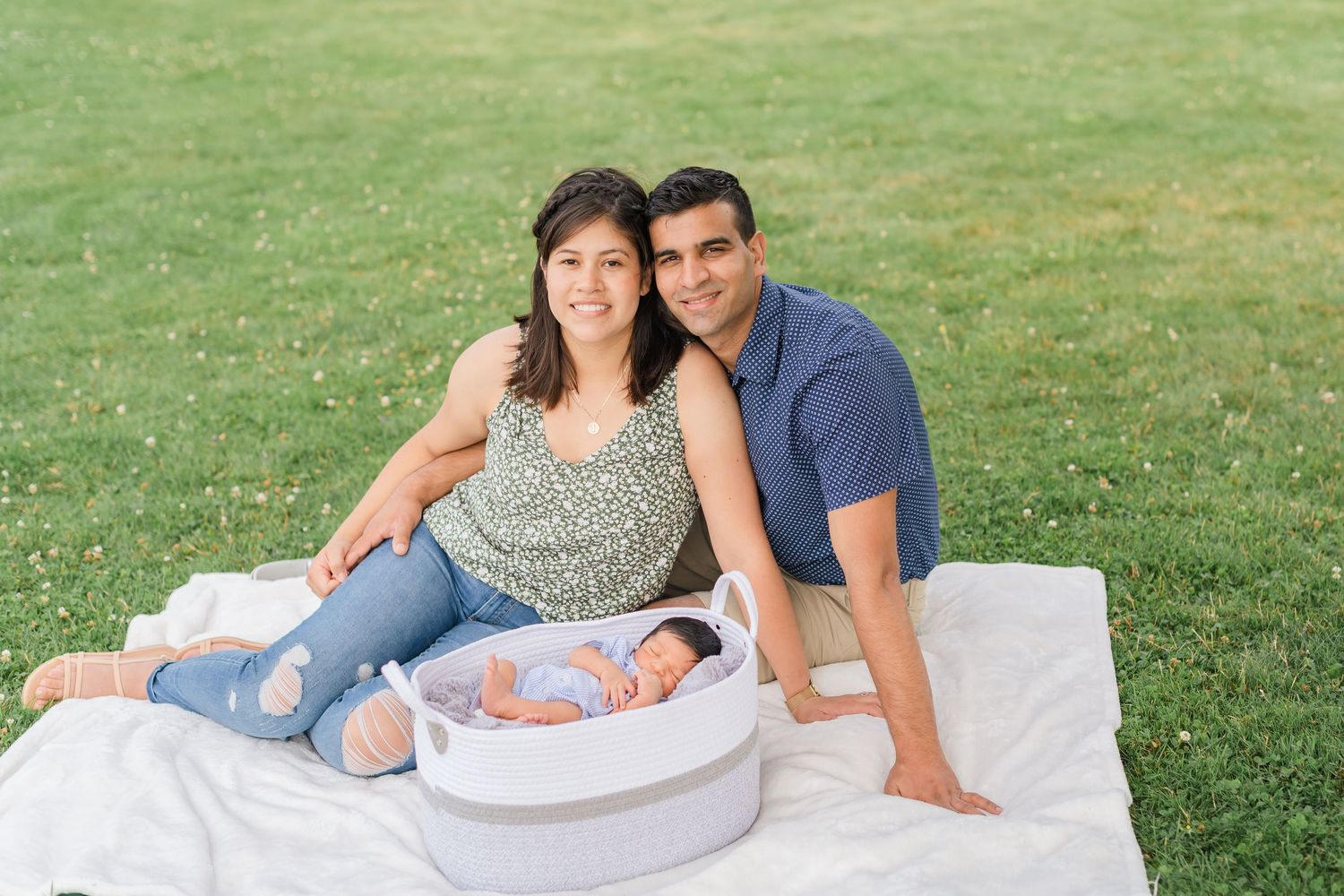 A family lies together on a white blanket in the grass during a summer outdoor photo session.