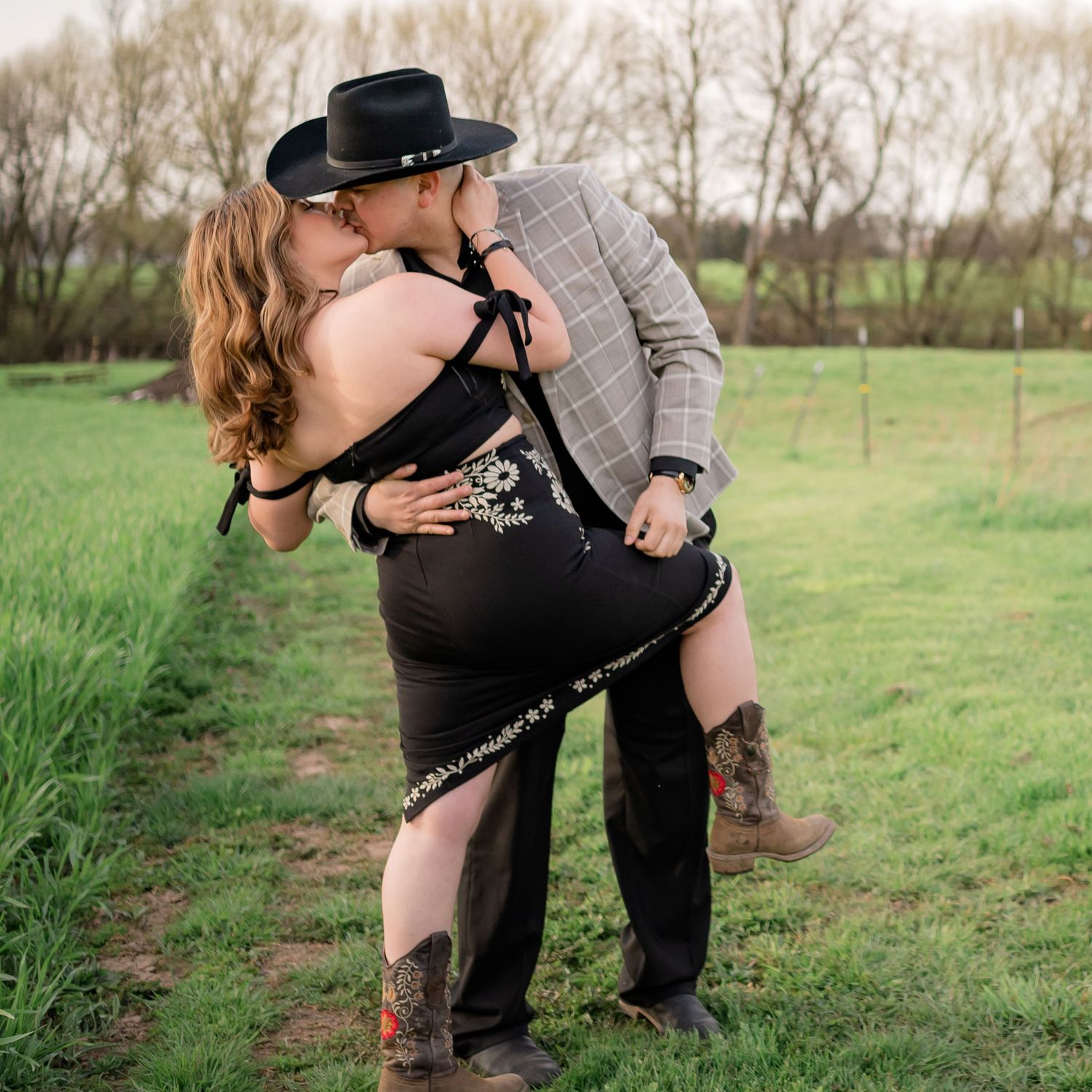 Silhouetted couple stands close together in a romantic embrace against a grassy field at sunset.