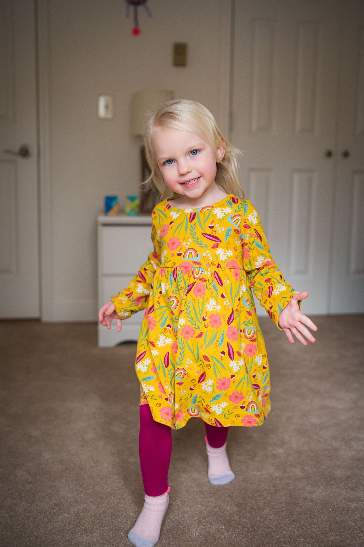 A series of playful dance poses in a yellow floral dress and burgundy leggings near white shelving.