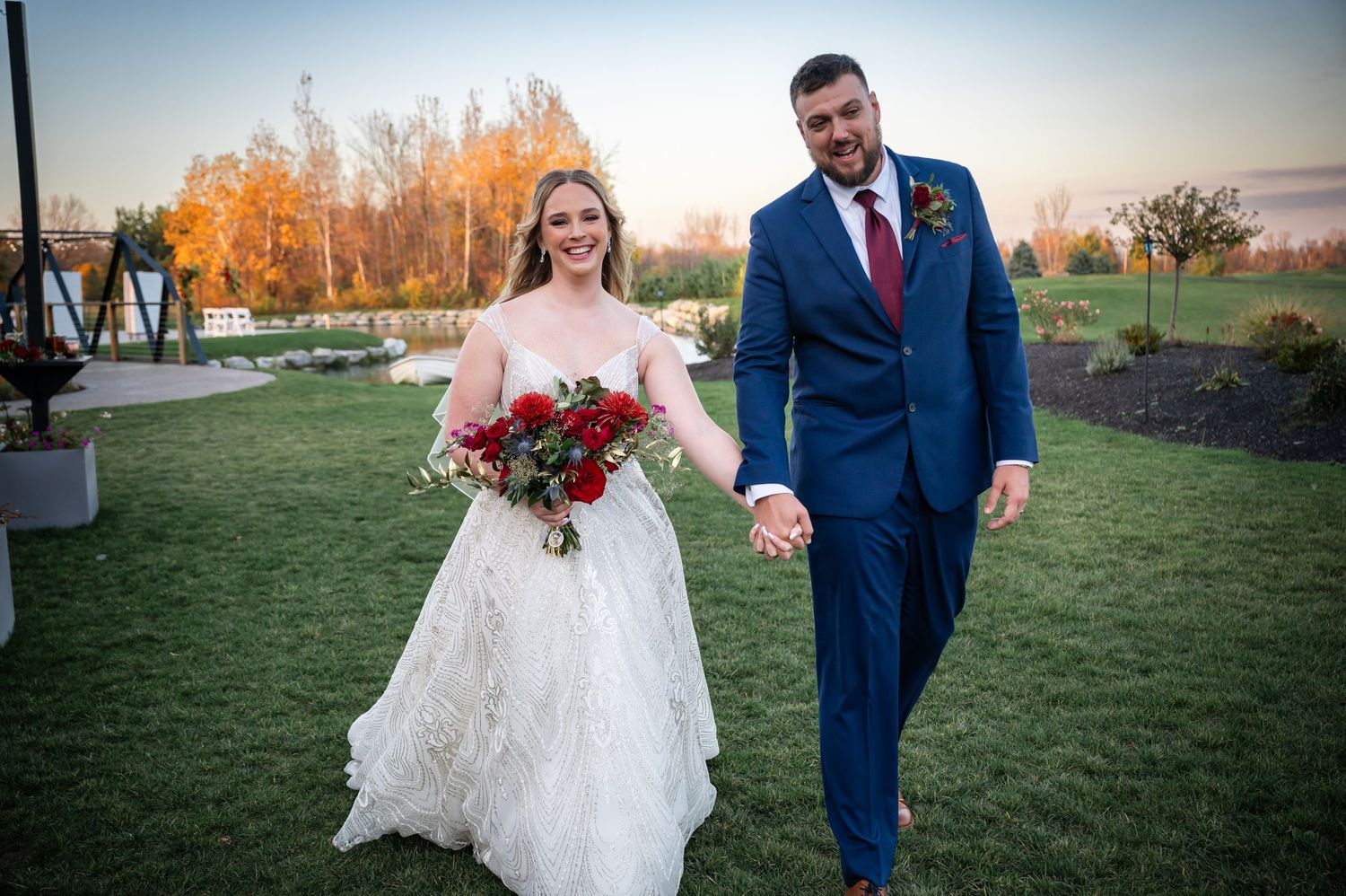 Newlyweds walking on grass with autumn colors and red bouquet at sunset.