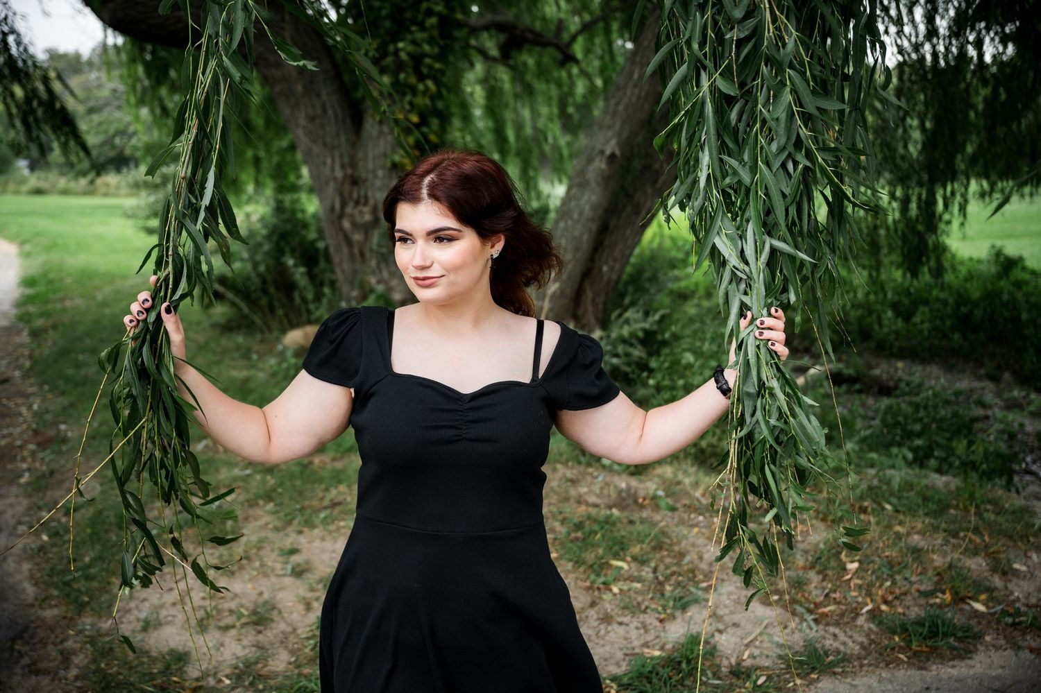 Figure in black dress stands between hanging willow branches in a garden setting.