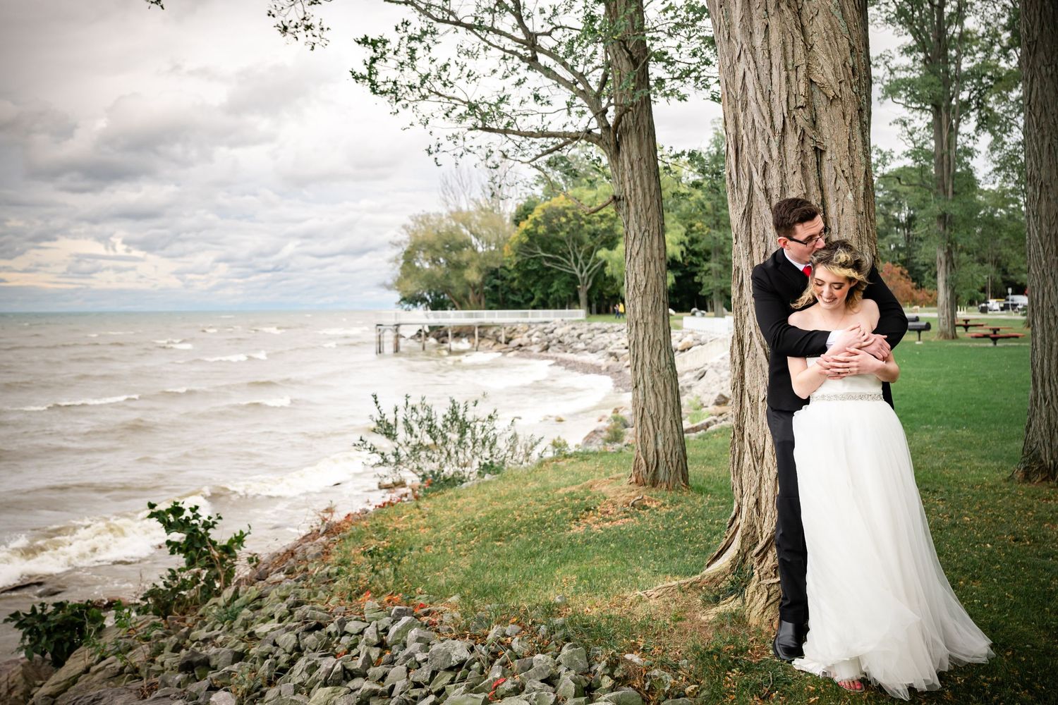 Couple embraces by large tree trunk with dramatic lake waves in background.