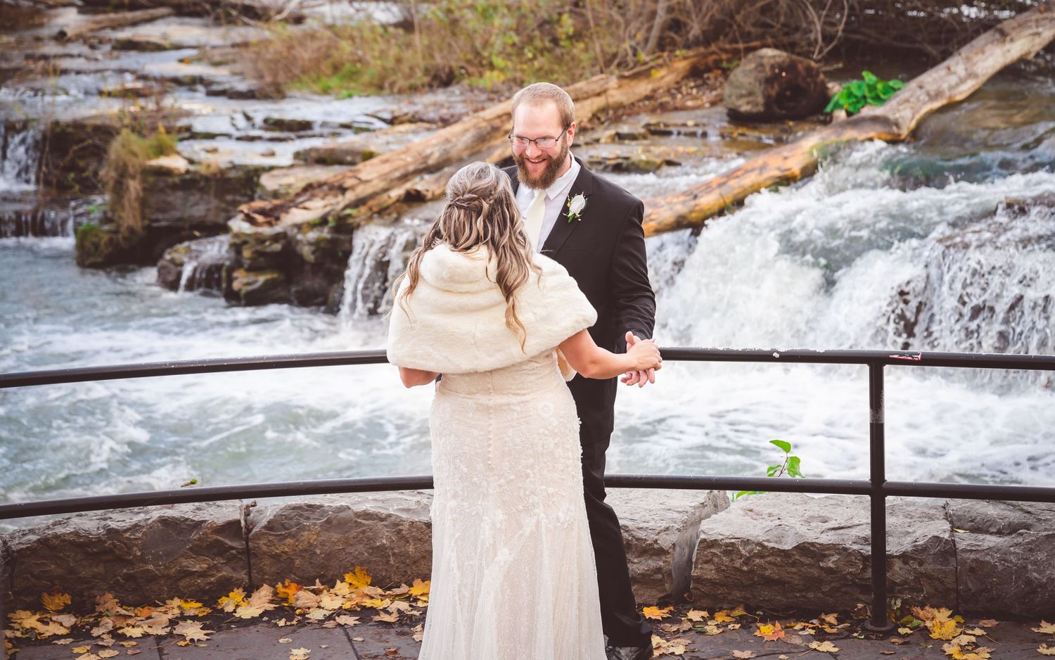 A wedding couple embraces near a scenic waterfall overlook with autumn leaves scattered on the ground.