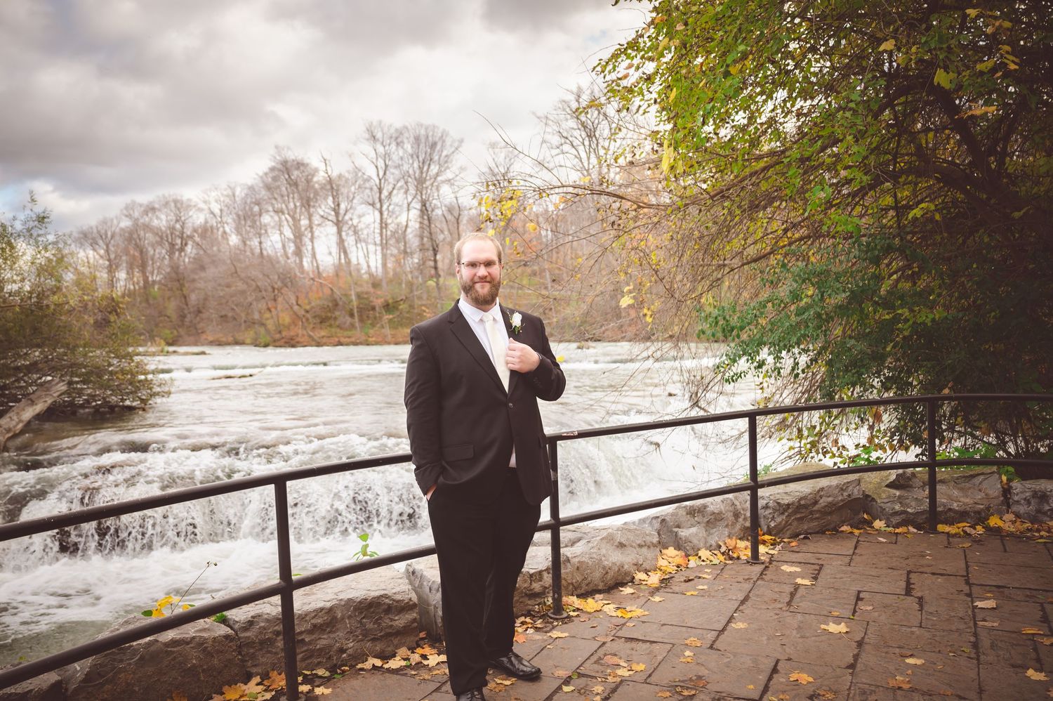 Person in formal attire stands on stone pathway overlooking a rushing river in autumn.