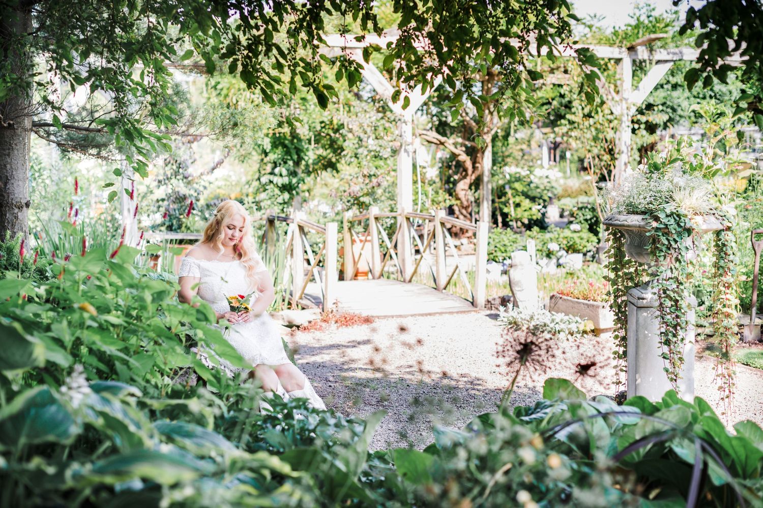 A white lace dress is visible through lush garden foliage on a sunny summer day.