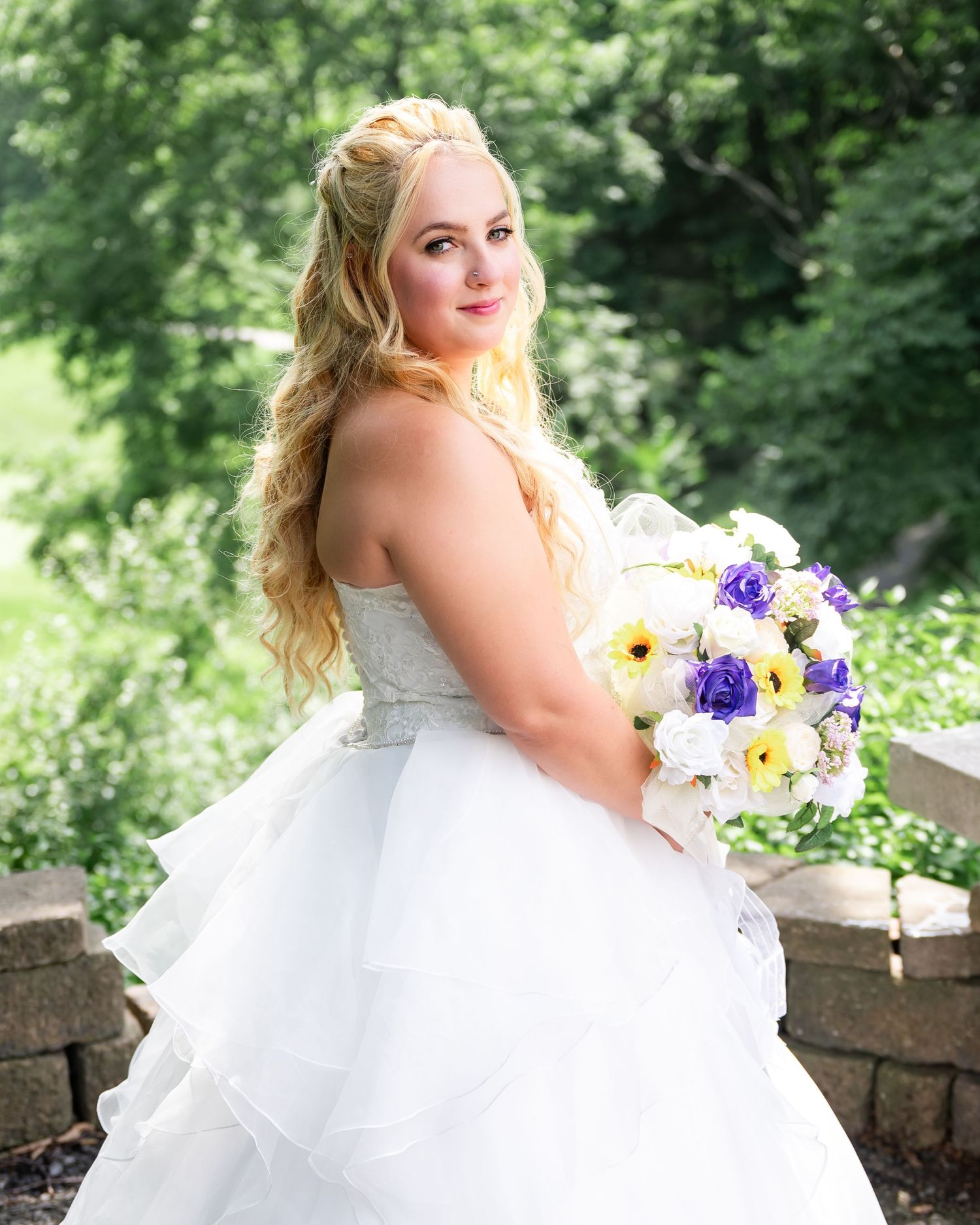 Bride in a flowing white dress poses with colorful bouquet on stone terrace with scenic backdrop.
