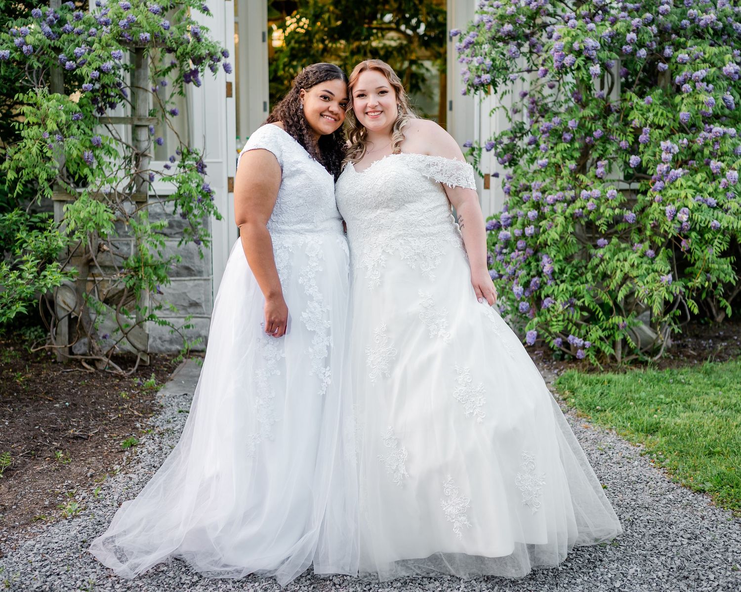 Couple in matching white wedding gowns pose in front of flower-covered garden archway.