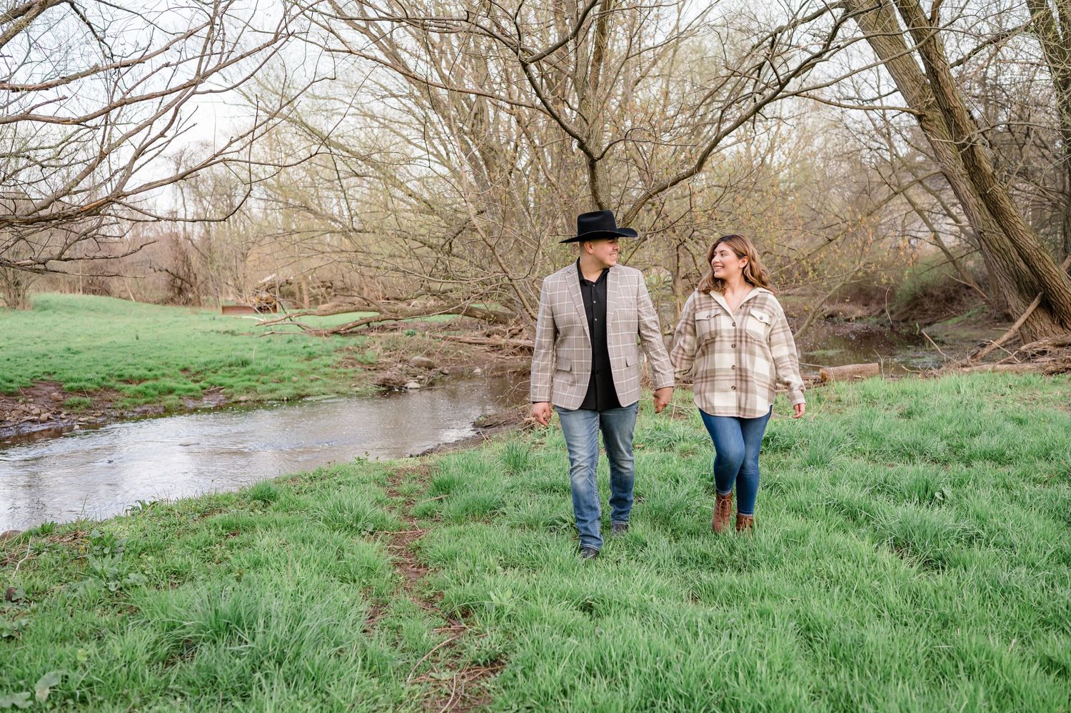 A couple takes a romantic stroll along a creek bank on lush green grass with bare trees in the background.