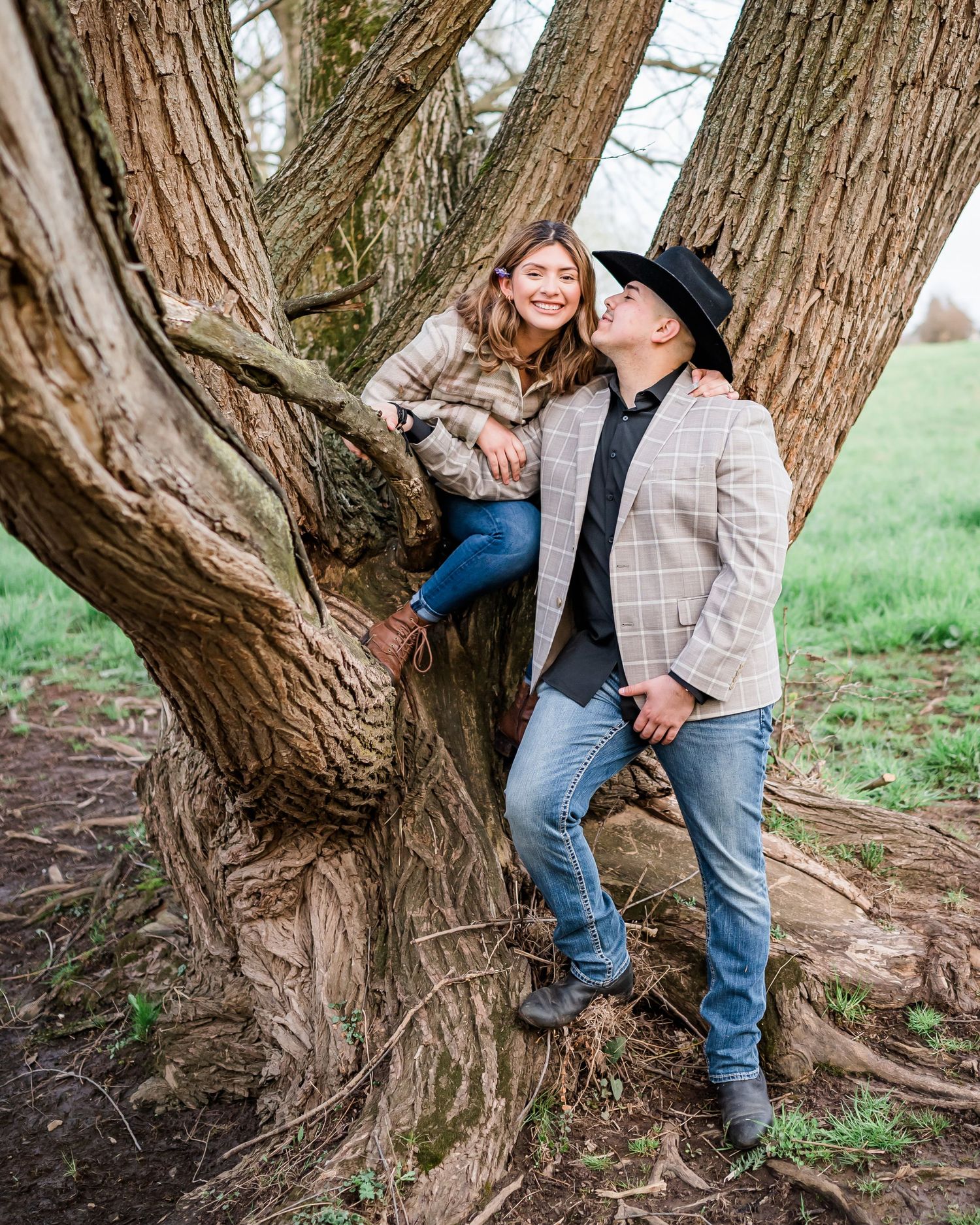 Playful couple poses among branches of large tree trunk in rural setting.