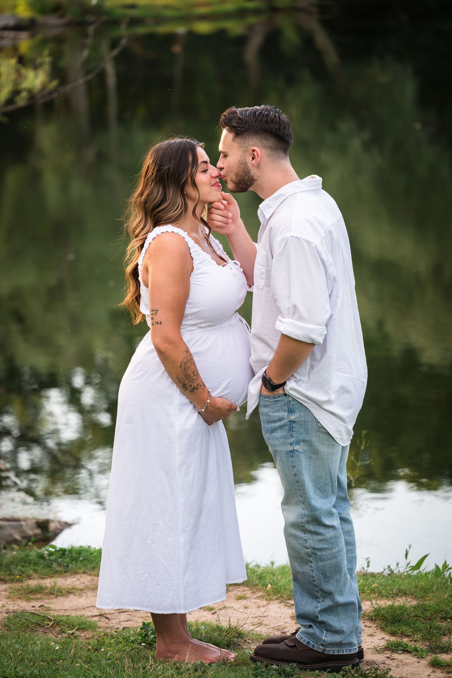 Pregnant couple sharing a tender moment by a serene pond, woman in white dress, man in white shirt and jeans.