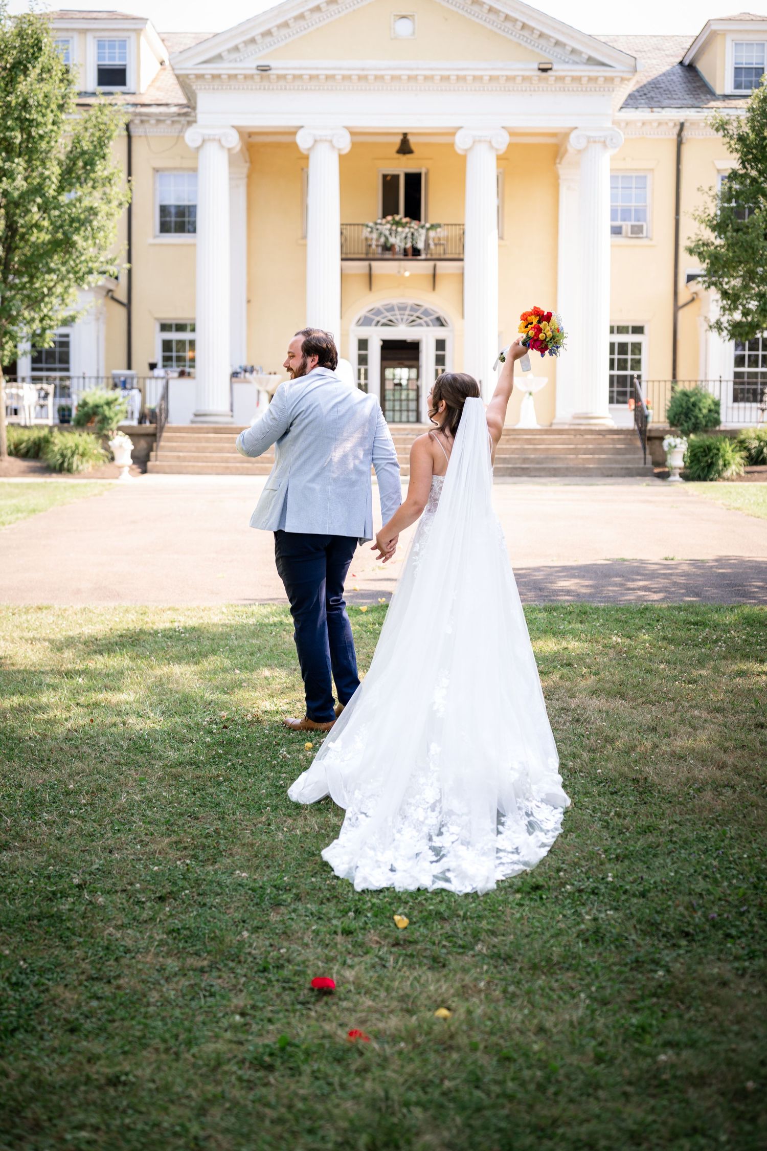 Bride in white gown holding bouquet walks with groom toward elegant yellow colonial mansion on wedding day.