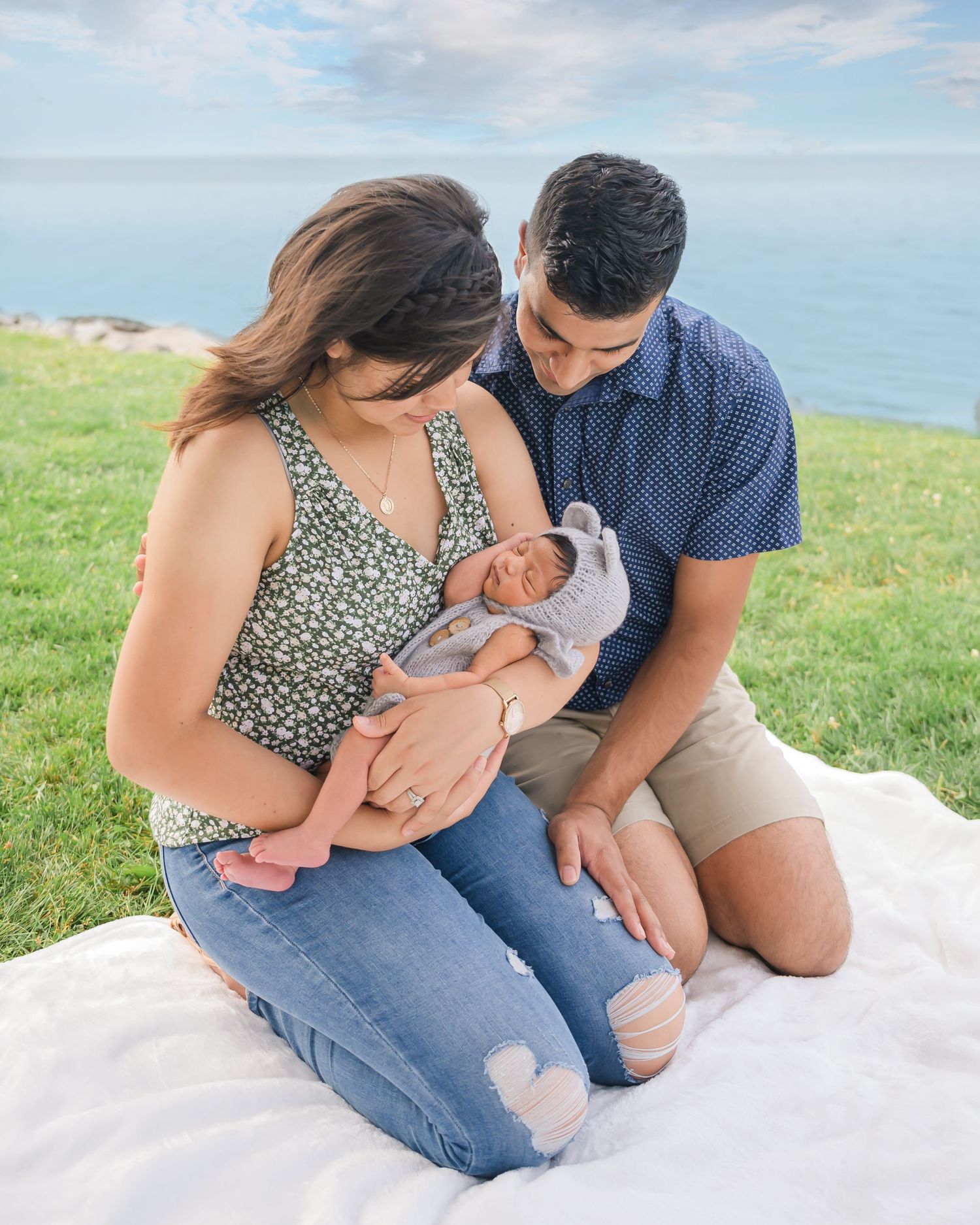 A couple sits on a white blanket on grassy coastal cliffs while tenderly holding their newborn baby.