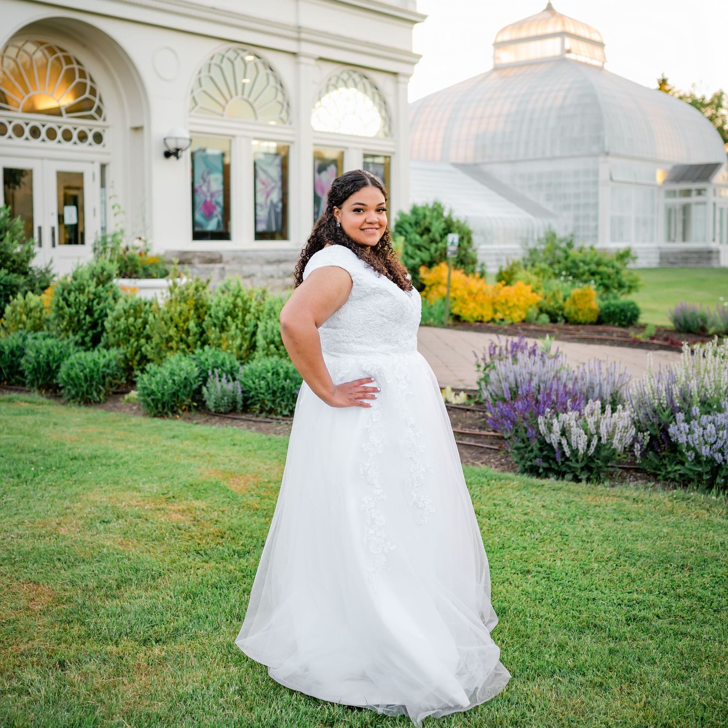 A bride poses in a white wedding gown against a classical building with garden landscaping.