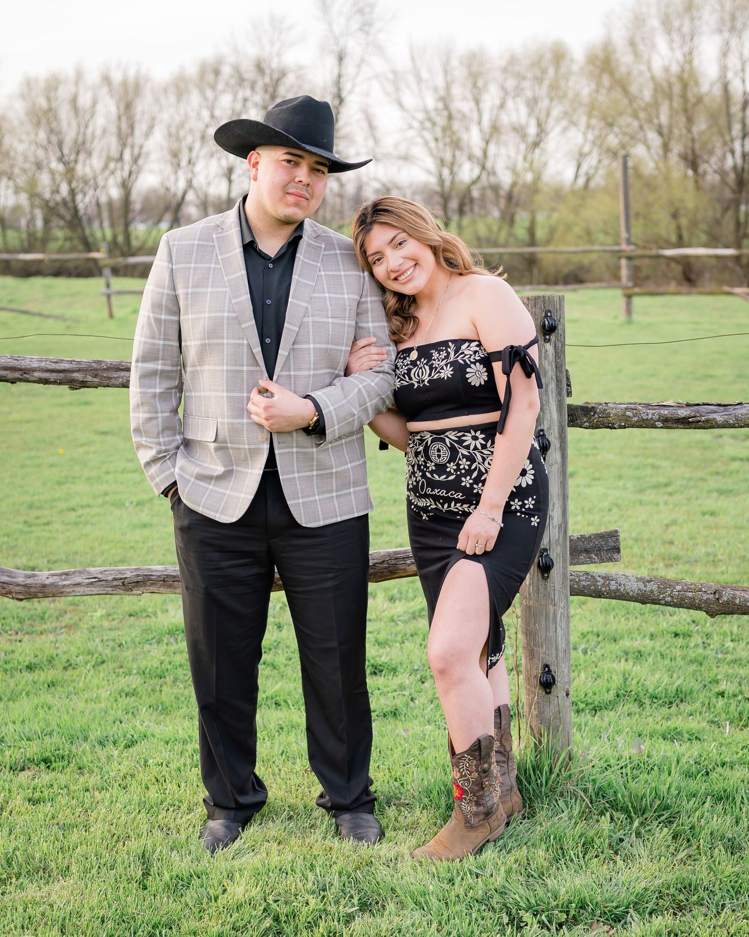 A couple poses casually against a wooden fence post in a country setting.