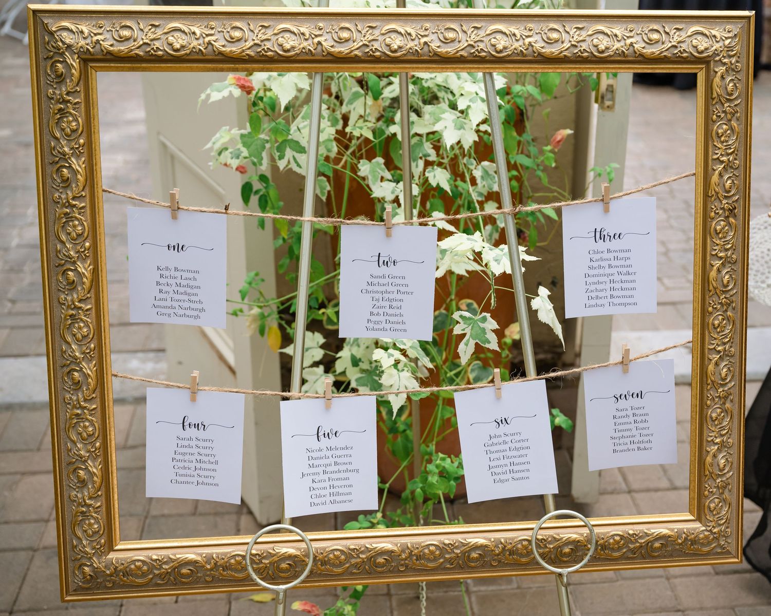 Gold ornate frame displaying wedding seating assignments on paper cards with floral backdrop.