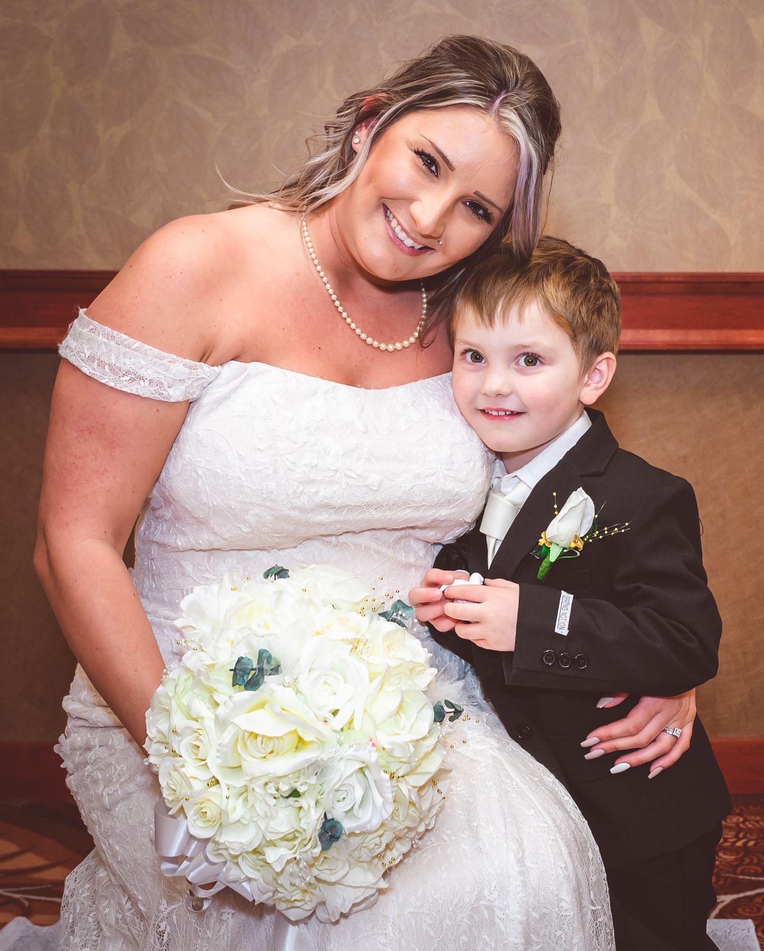 A bride in a white off-shoulder dress poses with a young ring bearer in a black suit holding a white bouquet.