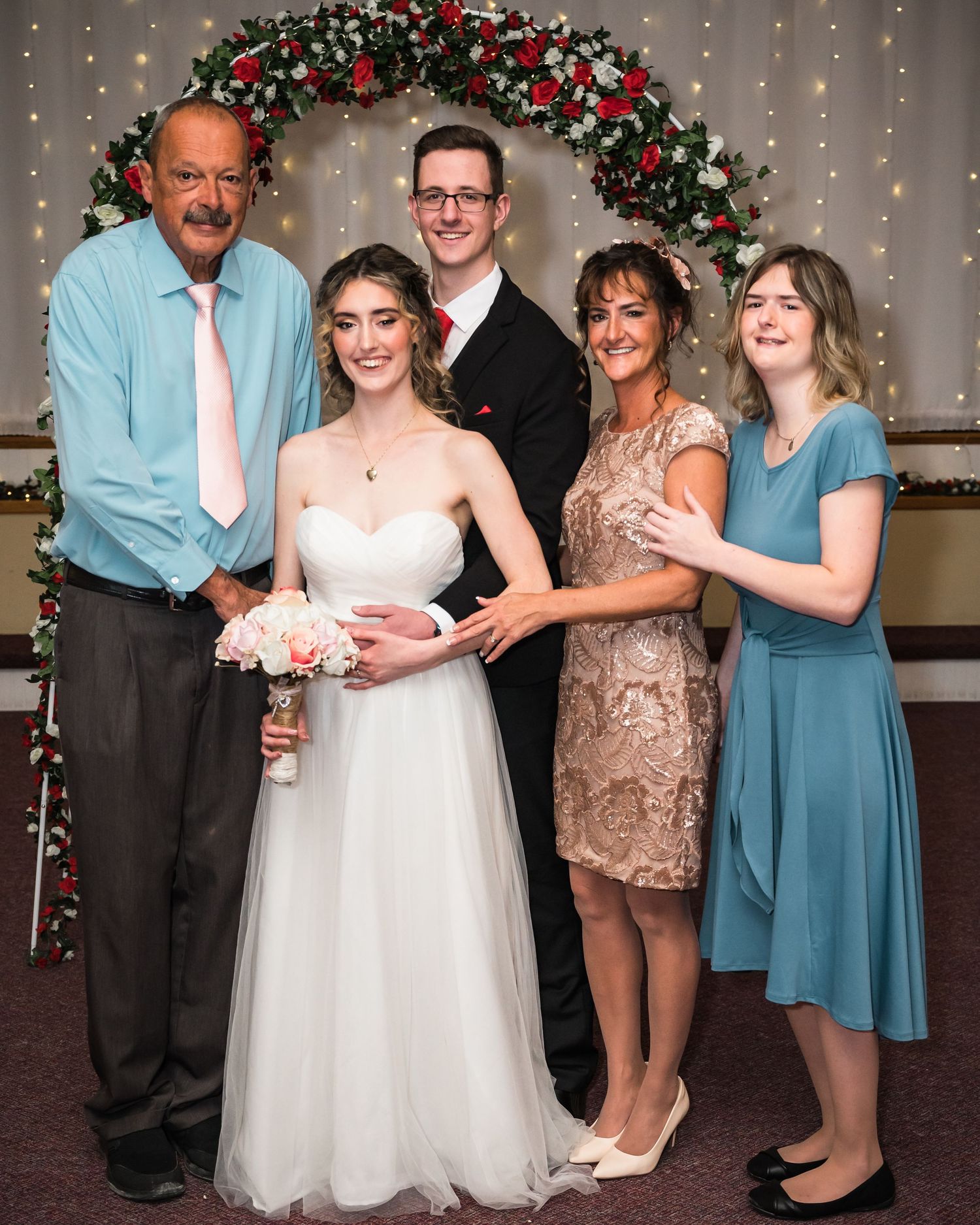 Family photo under an ornate Christmas arch with the bride in white and family members in formal attire.