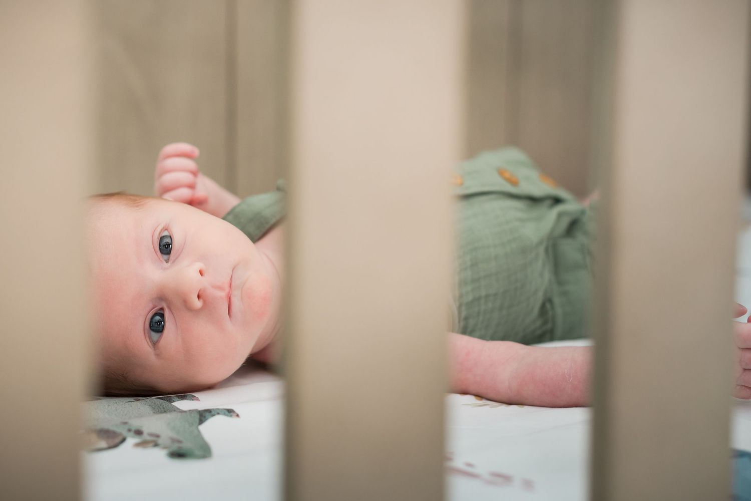A baby in a green outfit lies in a white crib looking up at the camera with curious eyes.