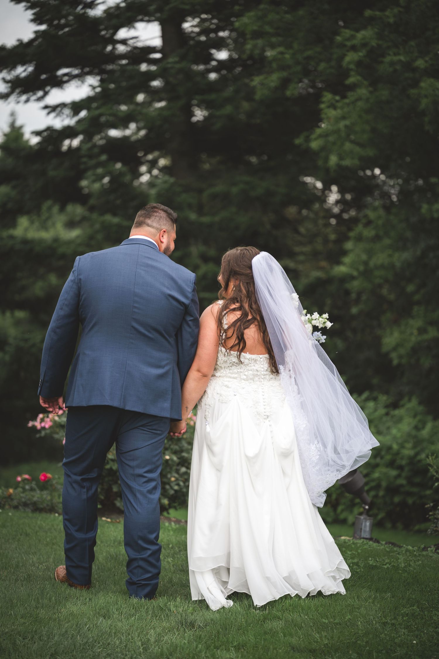 Bride and groom walking hand in hand through a lush garden, viewed from behind on their wedding day.
