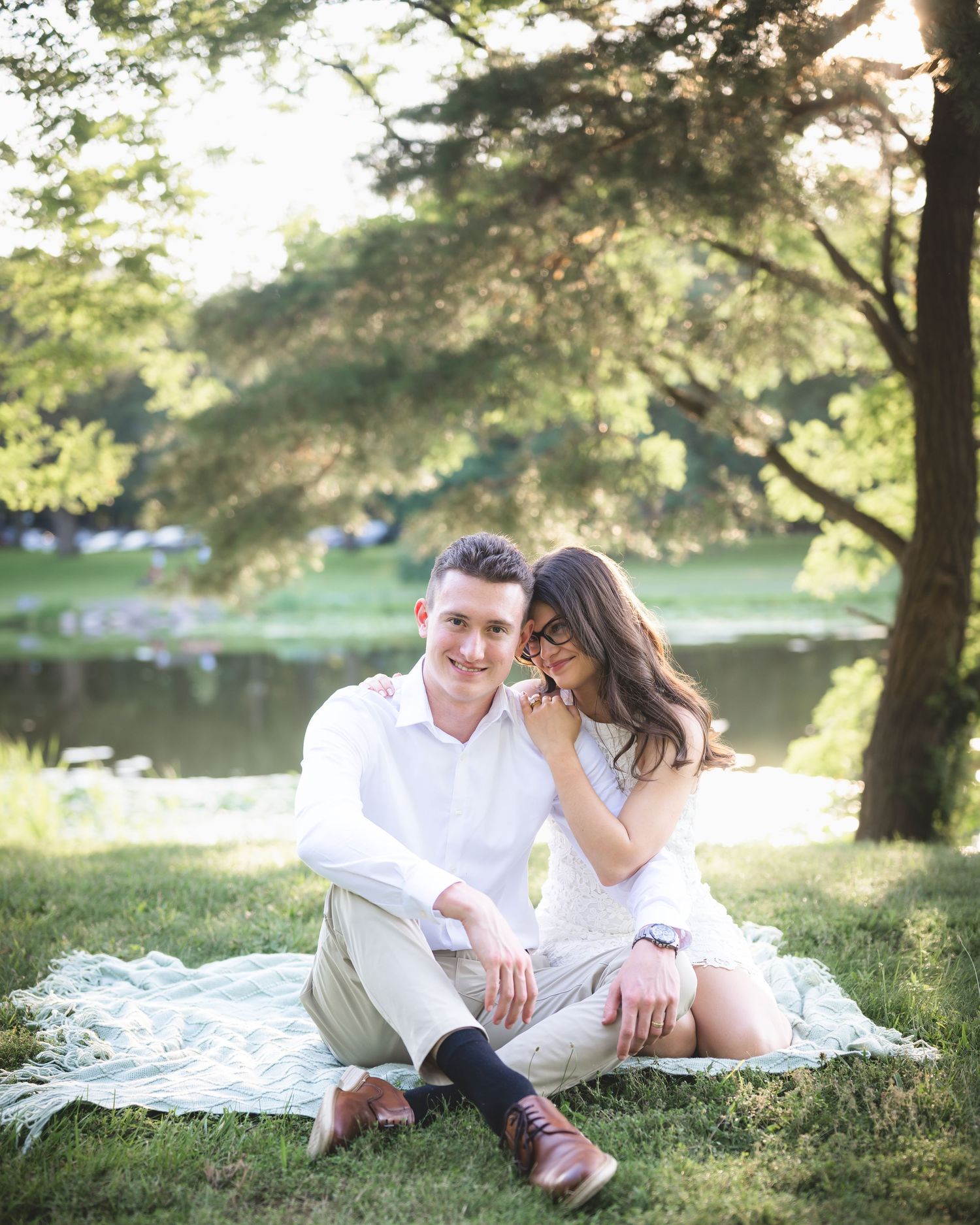 Couple sitting on blanket in park by pond, woman leaning on man's shoulder, both wearing white outfits.