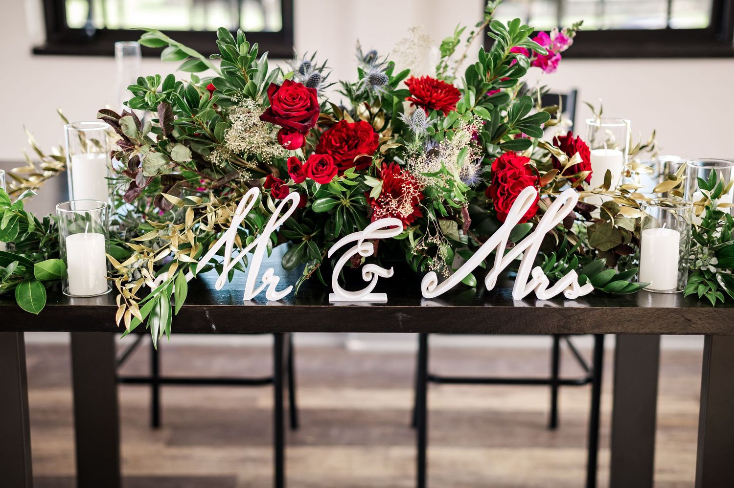 Mr & Mrs sign display with red floral arrangements and white candles on dark table.