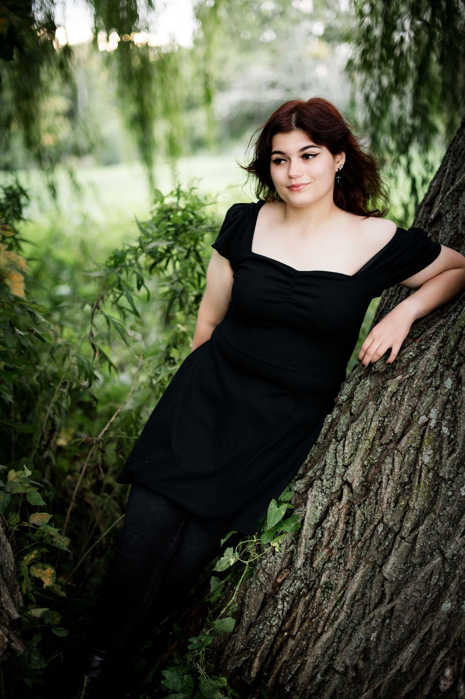 Portrait in black dress leaning against tree trunk with willow leaves in background.