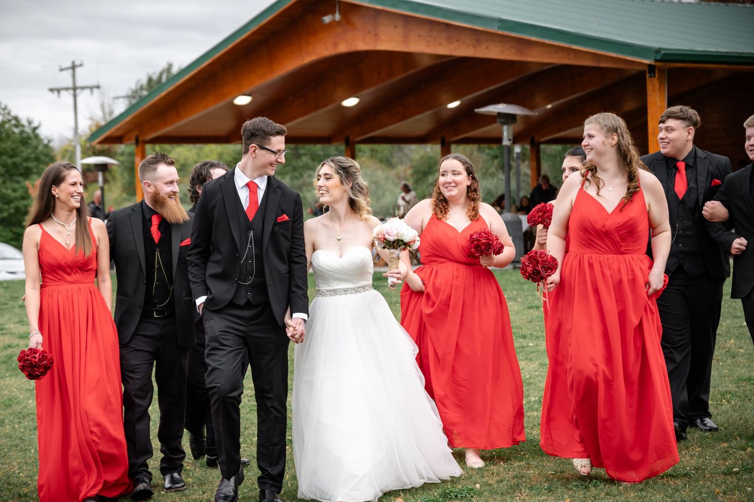 Wedding party in black suits and bright red dresses poses together under wooden pavilion.