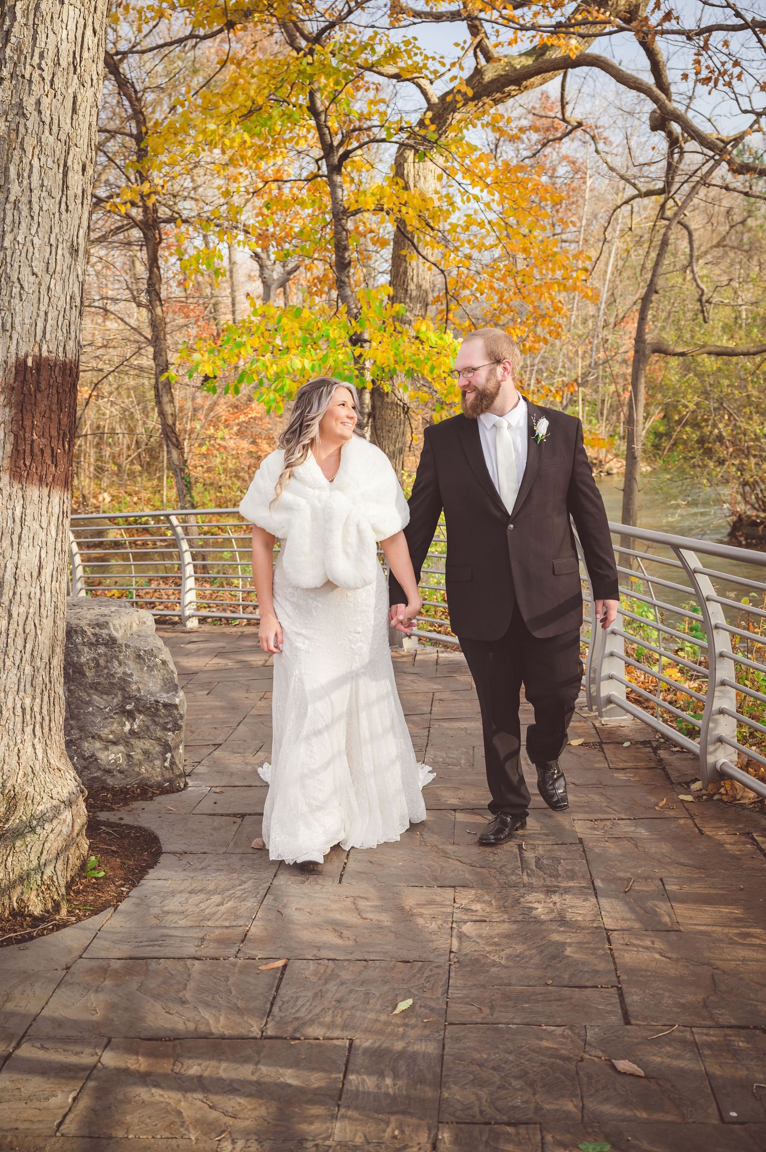 A couple walks hand in hand on a stone pathway surrounded by autumn trees and river views.