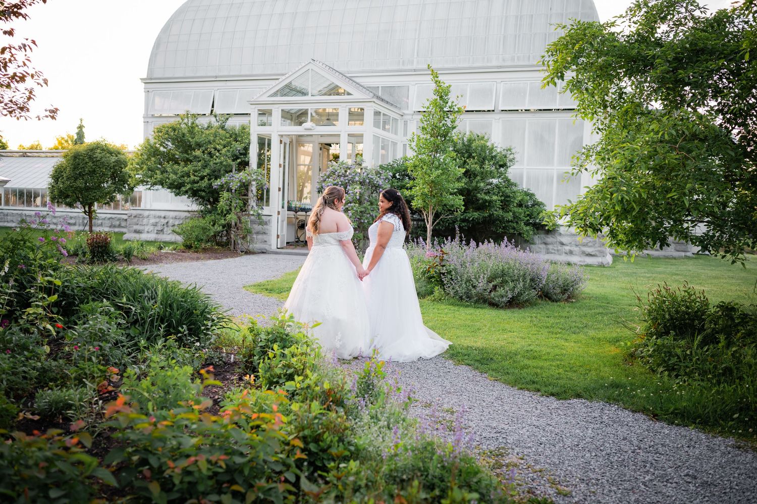 Two people in white wedding dresses walk along a garden path near a greenhouse structure.