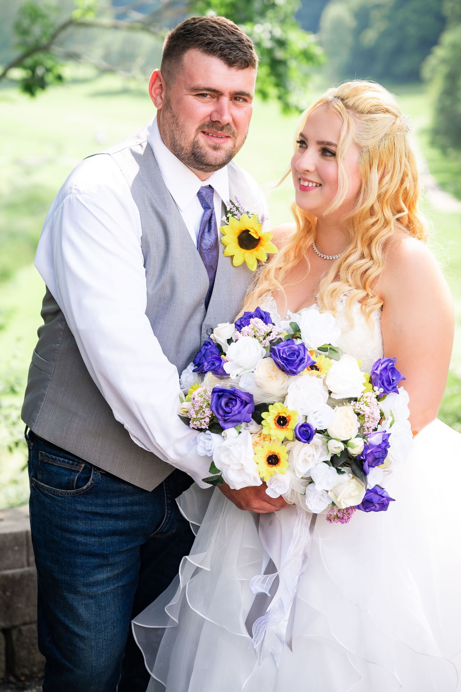 Wedding couple embracing on stone overlook with sunflower and purple rose bouquet.