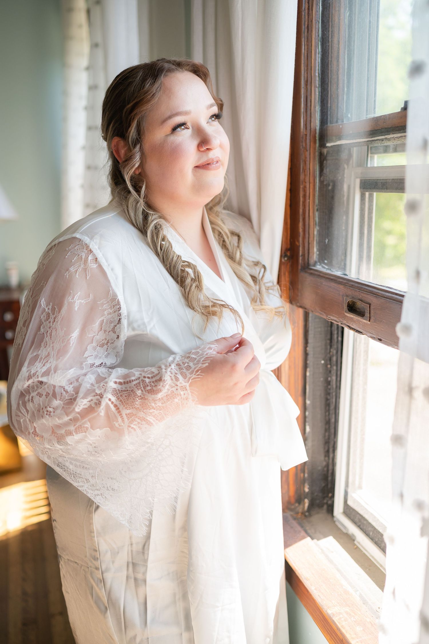 A person in a white lace robe gazes thoughtfully out a sunlit window.